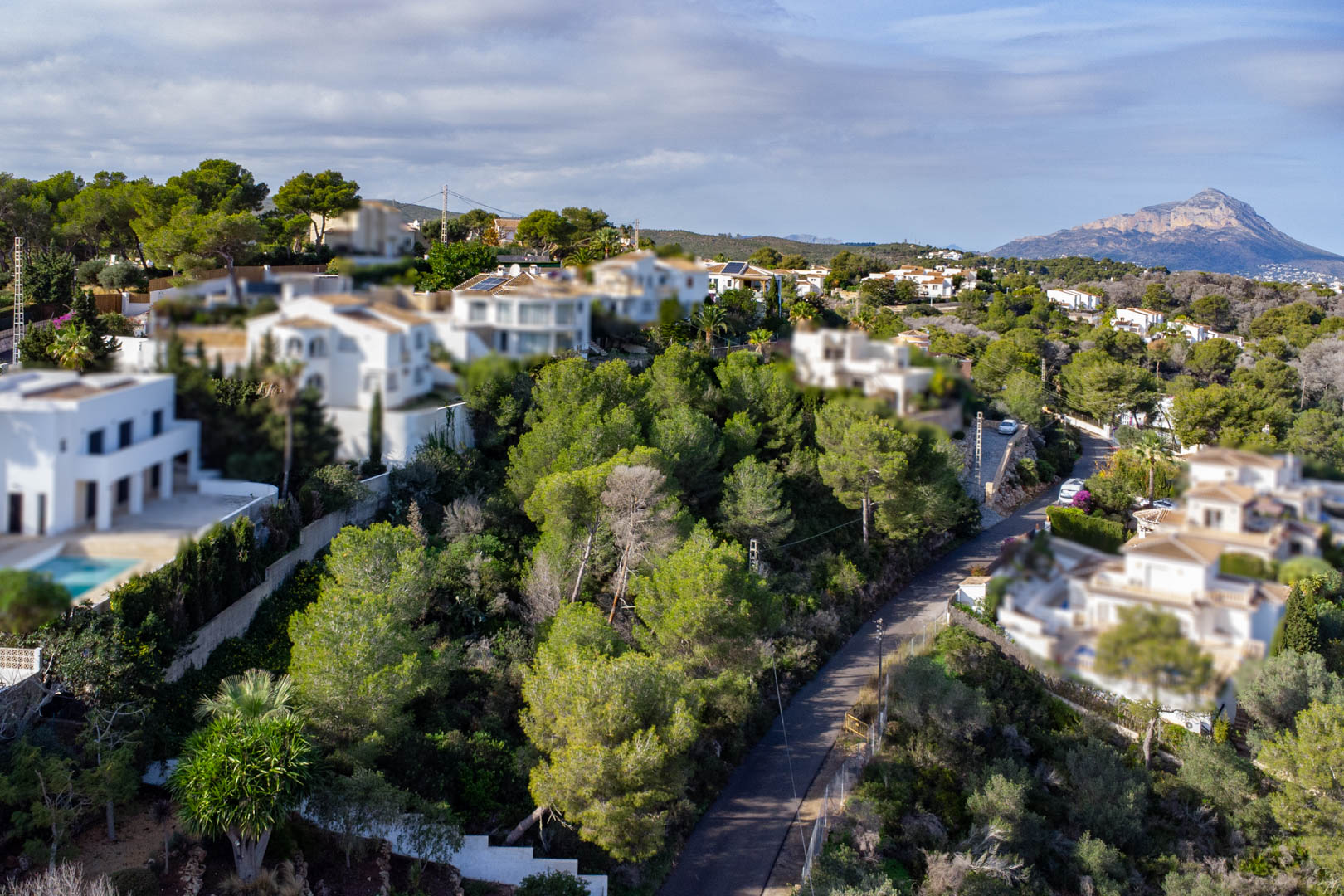 Vista aérea de la parcela arbolada en pendiente, rodeada de villas mediterráneas y con vistas lejanas al macizo del Montgó.