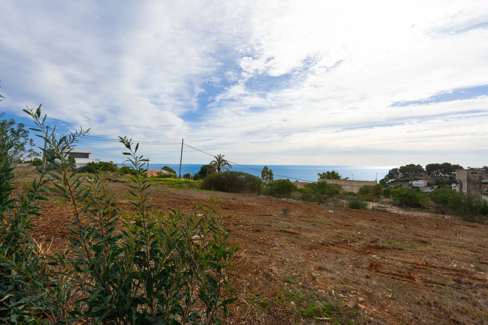 Parcela elevada con vistas panorámicas al mar, terreno despejado y vegetación mediterránea autóctona bajo un cielo parcialmente nublado.