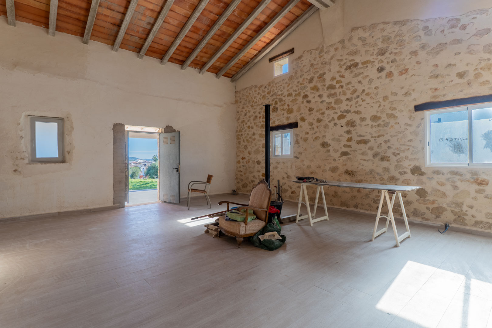 Estancia principal con muros de piedra seca original, techos altos con vigas de madera y baldosas de barro, y acceso directo al exterior con vistas despejadas.