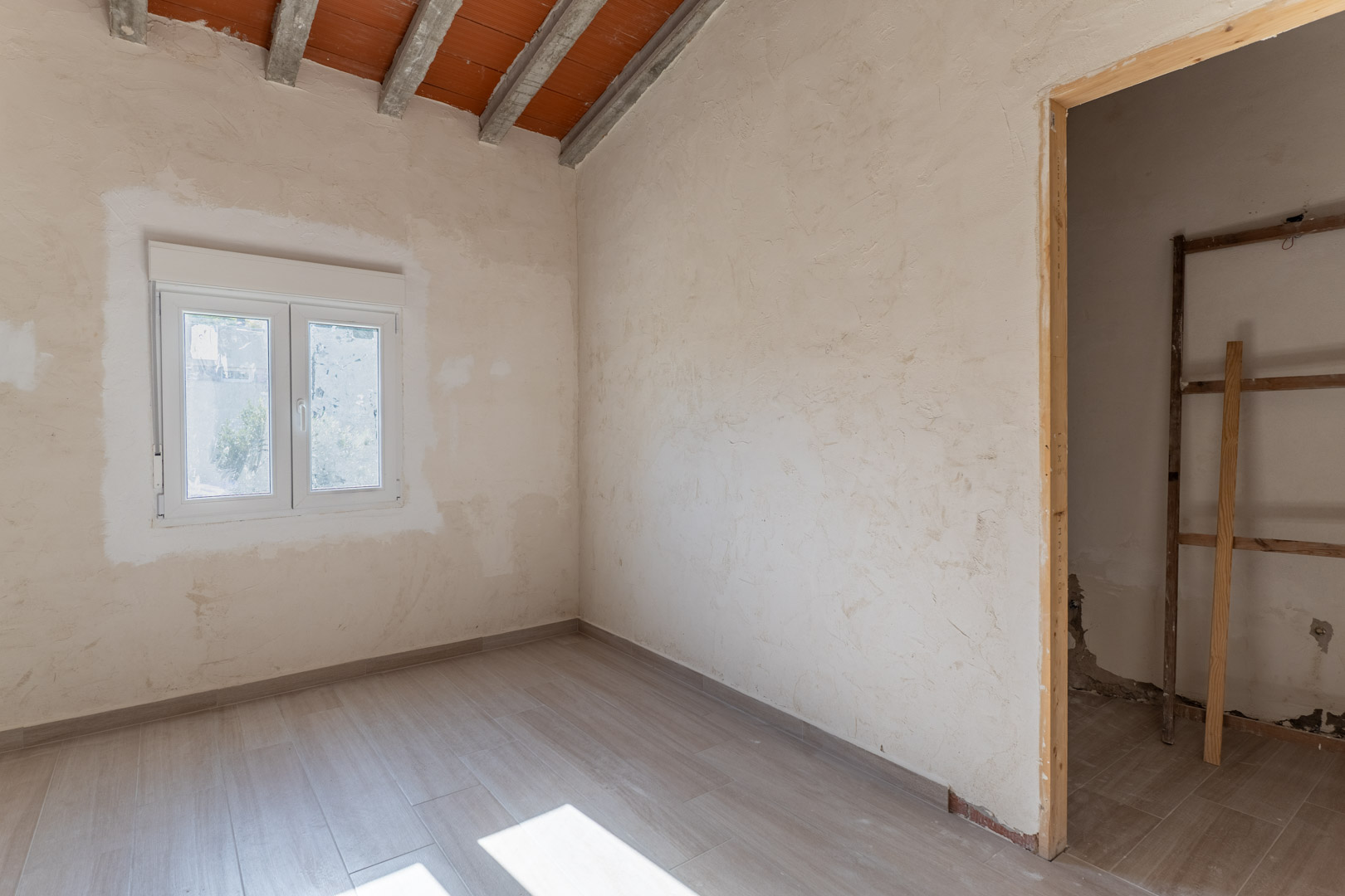 Habitación en reforma con suelos de gres imitación madera, ventana de PVC blanca y techo inclinado de vigas de hormigón con bovedillas vistas.