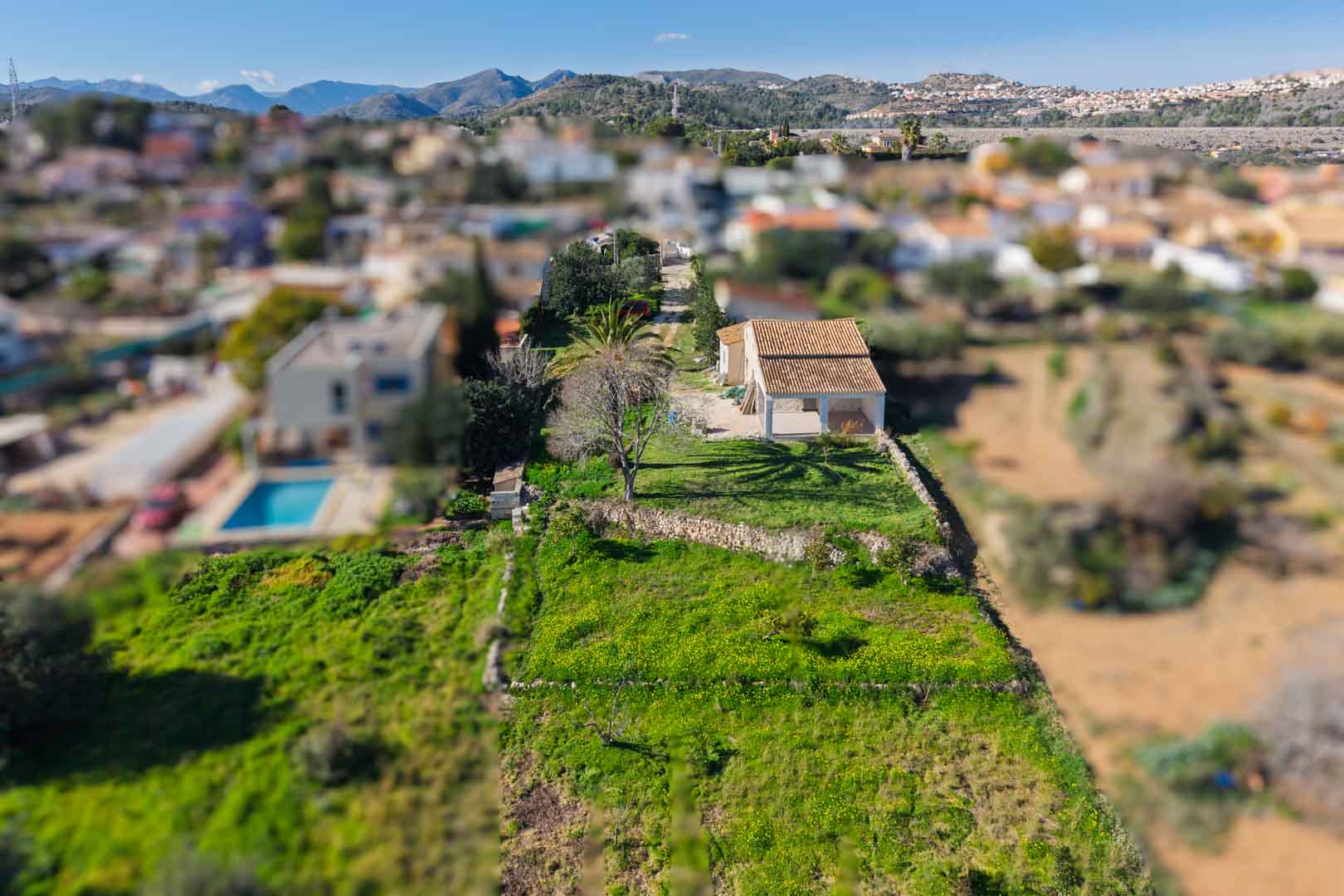 Vista aérea de la finca con su tejado renovado y naya, situada en una parcela alargada con muros de piedra seca y vistas panorámicas a las montañas circundantes.
