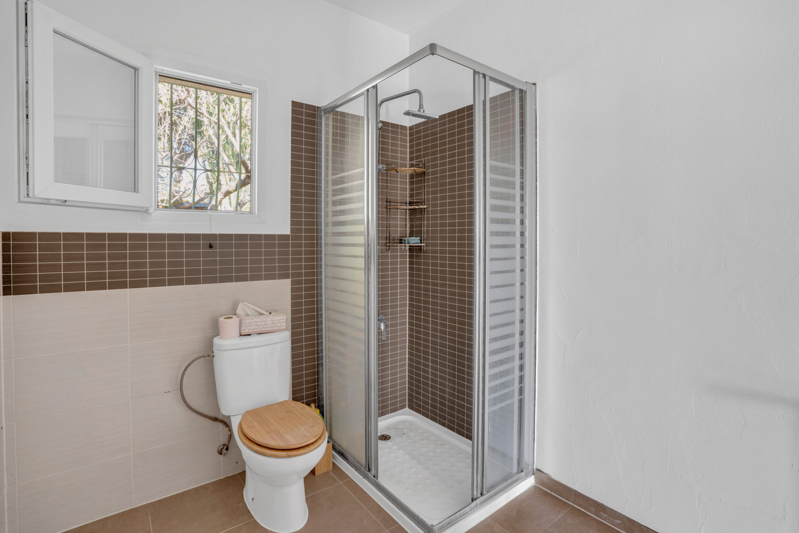 Cuarto de baño funcional con ducha de rincón revestida en gresite marrón, mampara de cristal, inodoro con tapa de madera y ventana para ventilación natural.