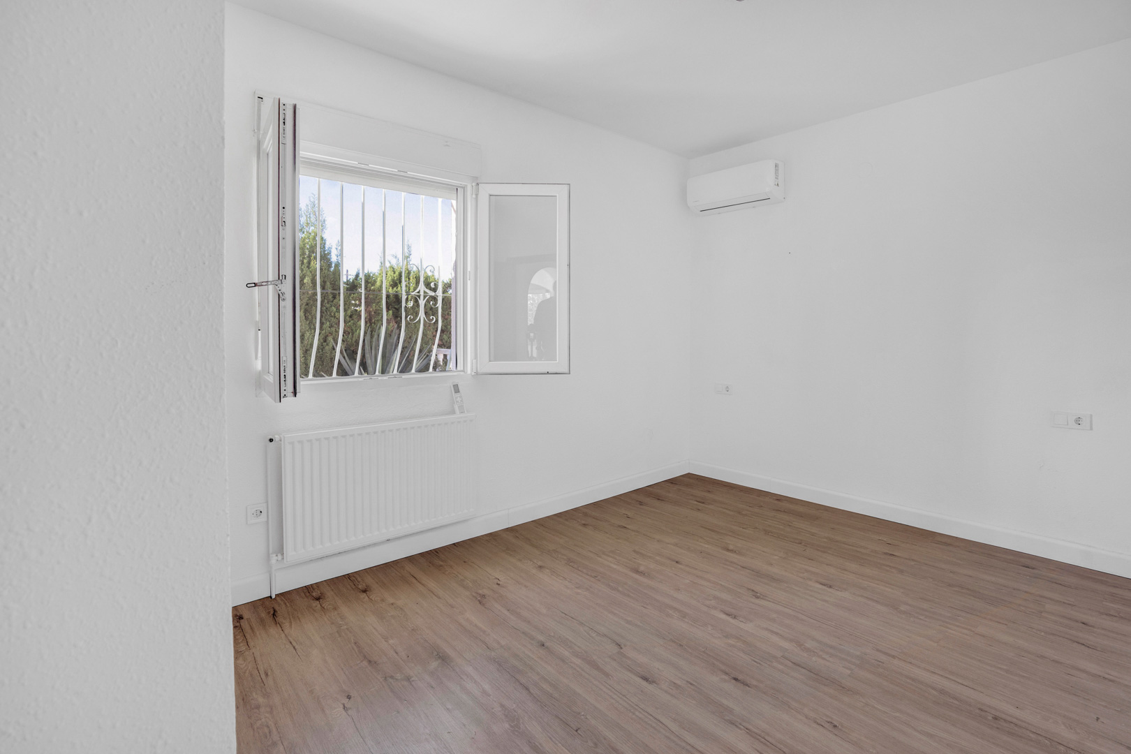 Dormitorio luminoso con suelos de tarima laminada, equipado con radiador de calefacción central y aire acondicionado split sobre paredes blancas.