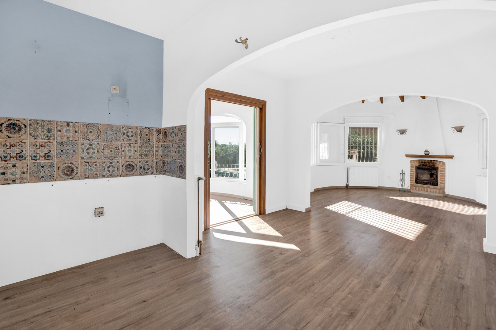 Salón-comedor diáfano con chimenea de obra, suelos de tarima y zona de cocina con azulejos decorativos. Los arcos conectan con una luminosa terraza acristalada.