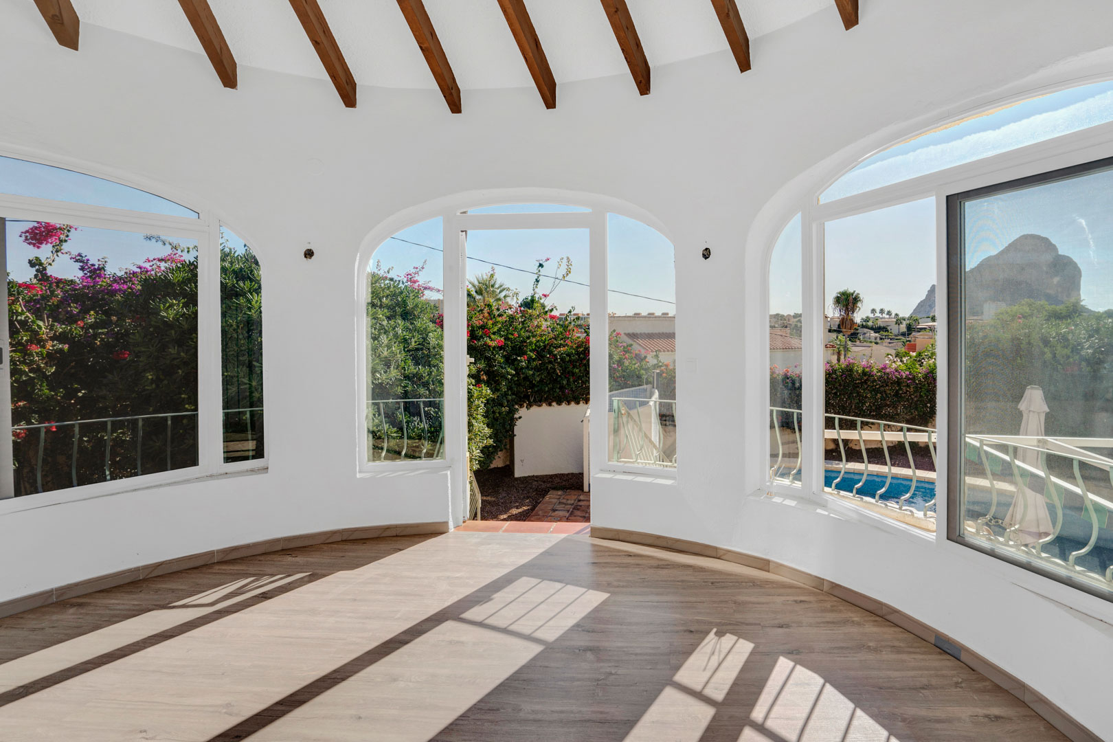 Terraza acristalada con vigas de madera vistas, suelos laminados y ventanales arqueados que ofrecen vistas directas al Peñón de Ifach y a la zona de la piscina.