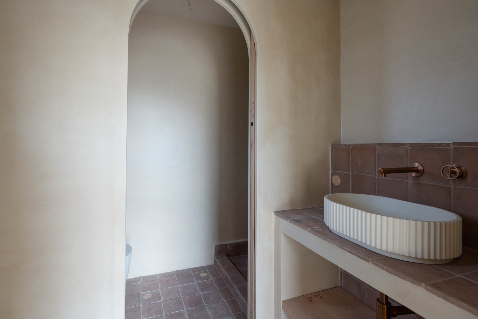Baño con paredes de arcilla, lavabo sobre encimera estriado y grifería empotrada en bronce. El diseño destaca por su arco de paso y suelos de terracota natural.