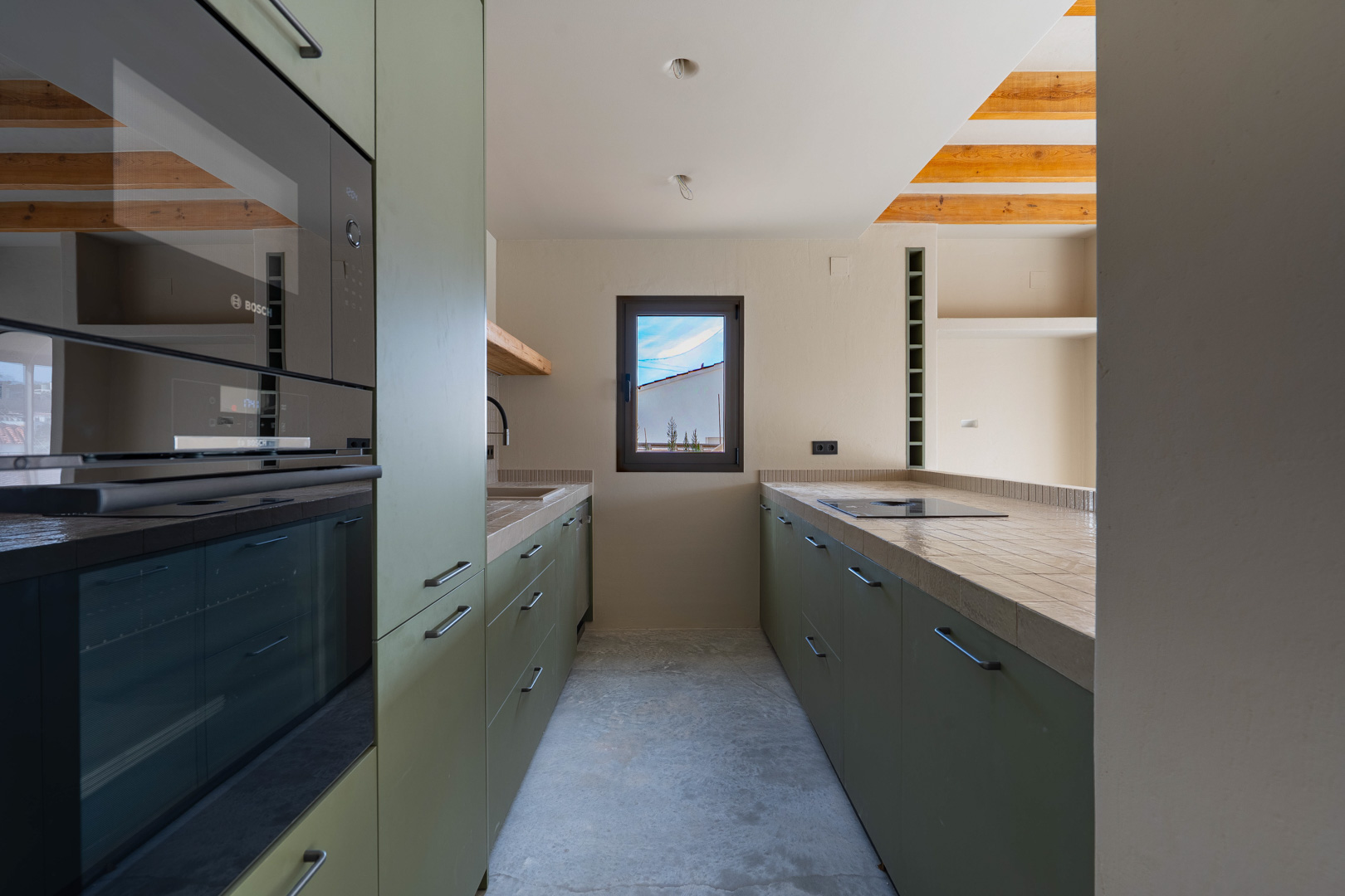 Cocina de diseño con armarios verde oliva, electrodomésticos Bosch integrados y encimeras porcelánicas. Destacan las vigas de madera vista y el suelo de hormigón pulido.