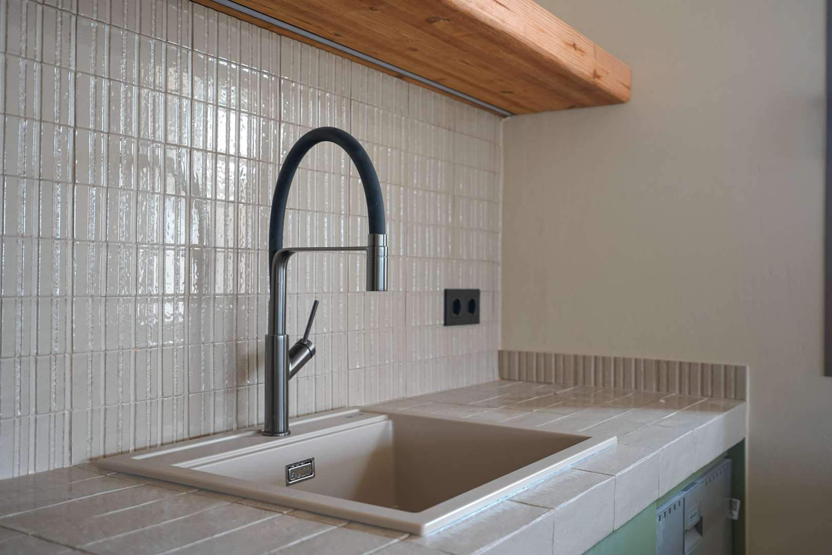 Detalle de cocina con encimera de gres porcelánico y fregadero integrado. Grifería moderna de cuello flexible sobre salpicadero de mosaico vertical y estante de madera maciza.