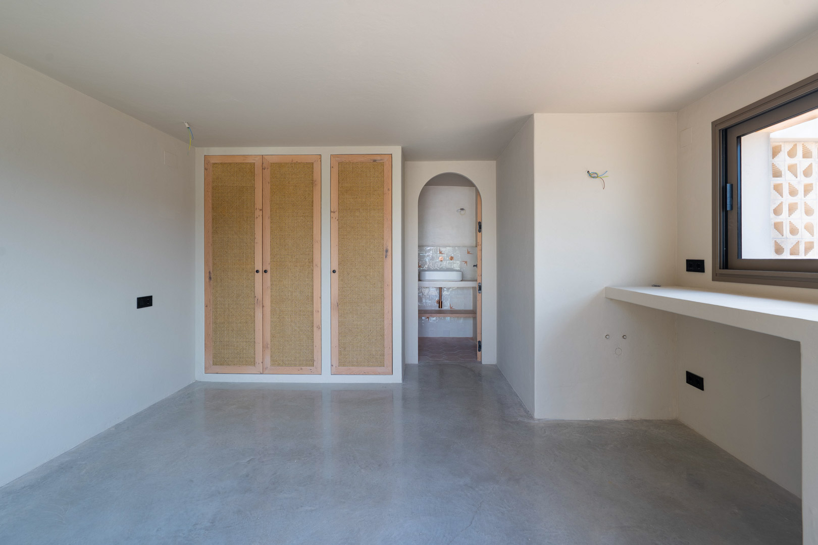 Dormitorio con armarios empotrados de roble y rejilla, suelos de hormigón pulido y acceso mediante arco a baño en suite con acabados de arcilla.