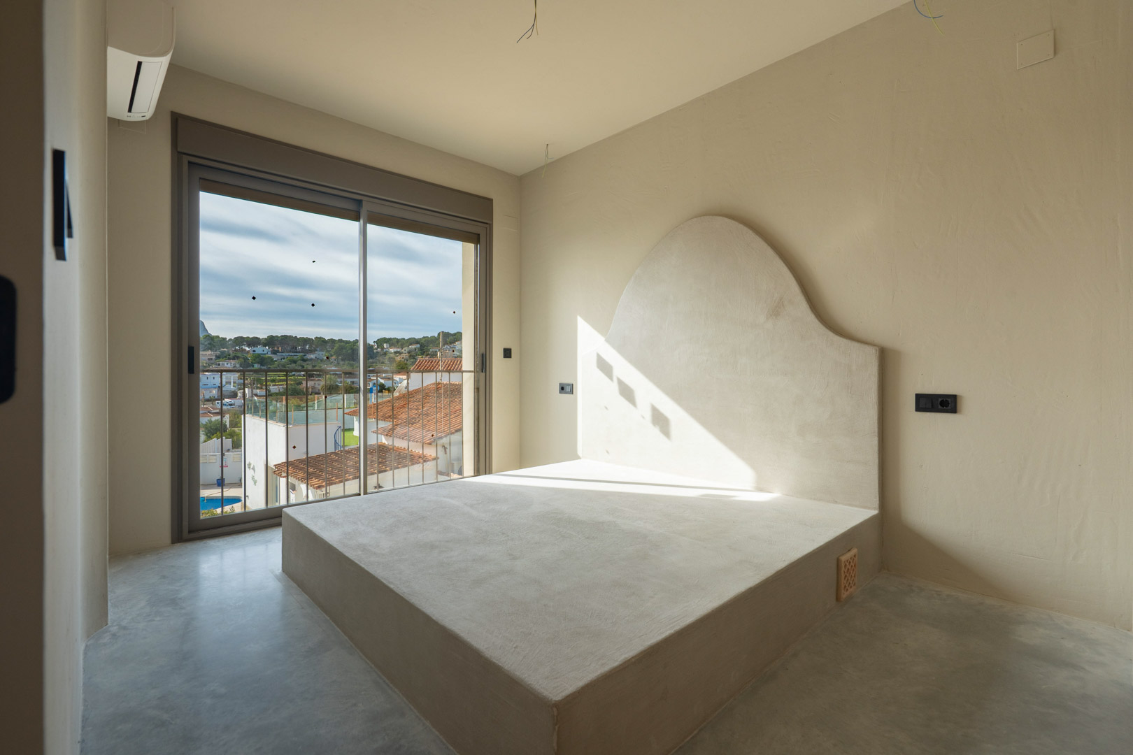 Dormitorio con base de cama integrada y cabecero curvo en acabado de arcilla, suelo de hormigón pulido y acceso a balcón con vistas despejadas.