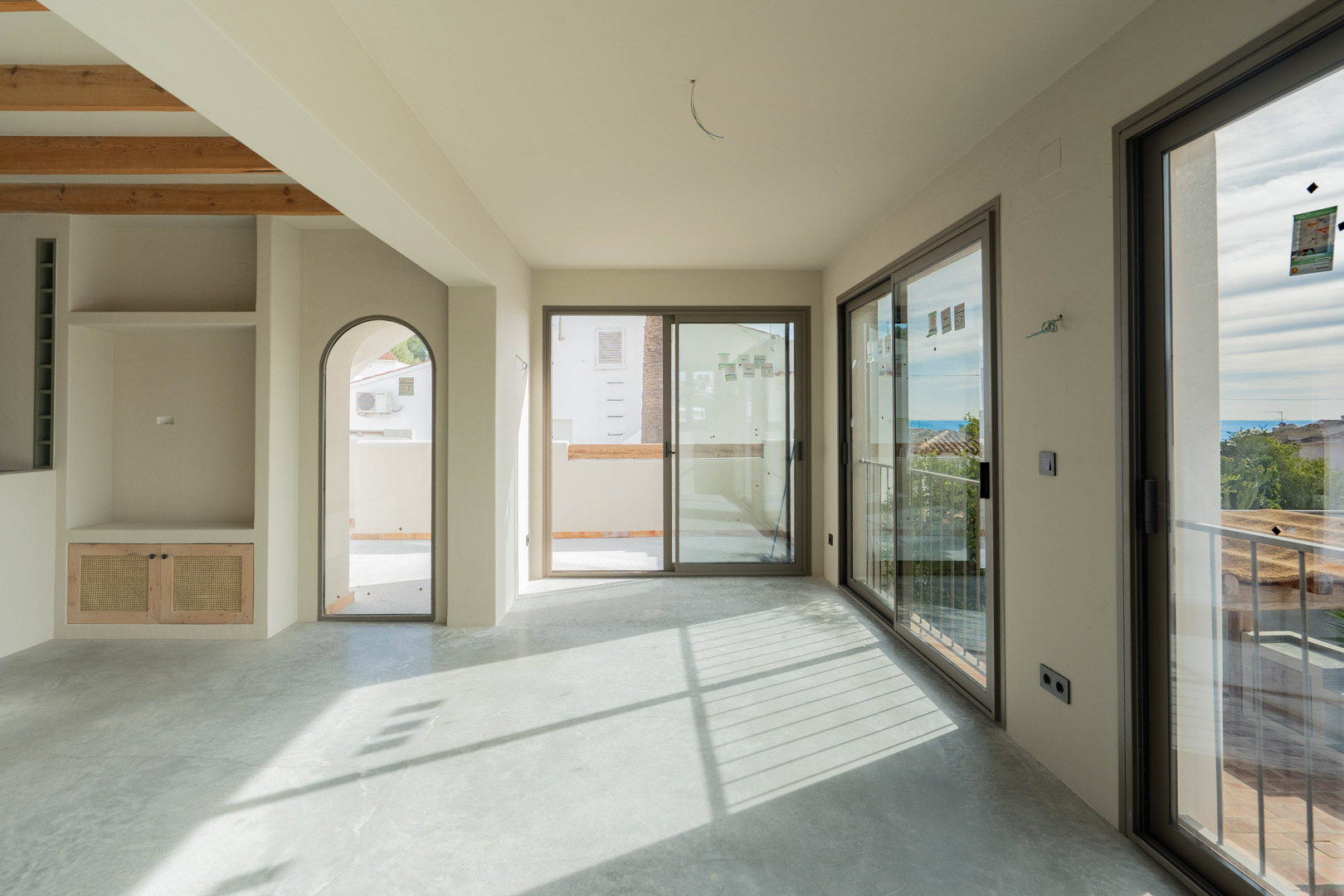Espacioso salón con suelos de hormigón pulido, estantería integrada y amplios ventanales correderos que capturan la luz natural y vistas al Mediterráneo.