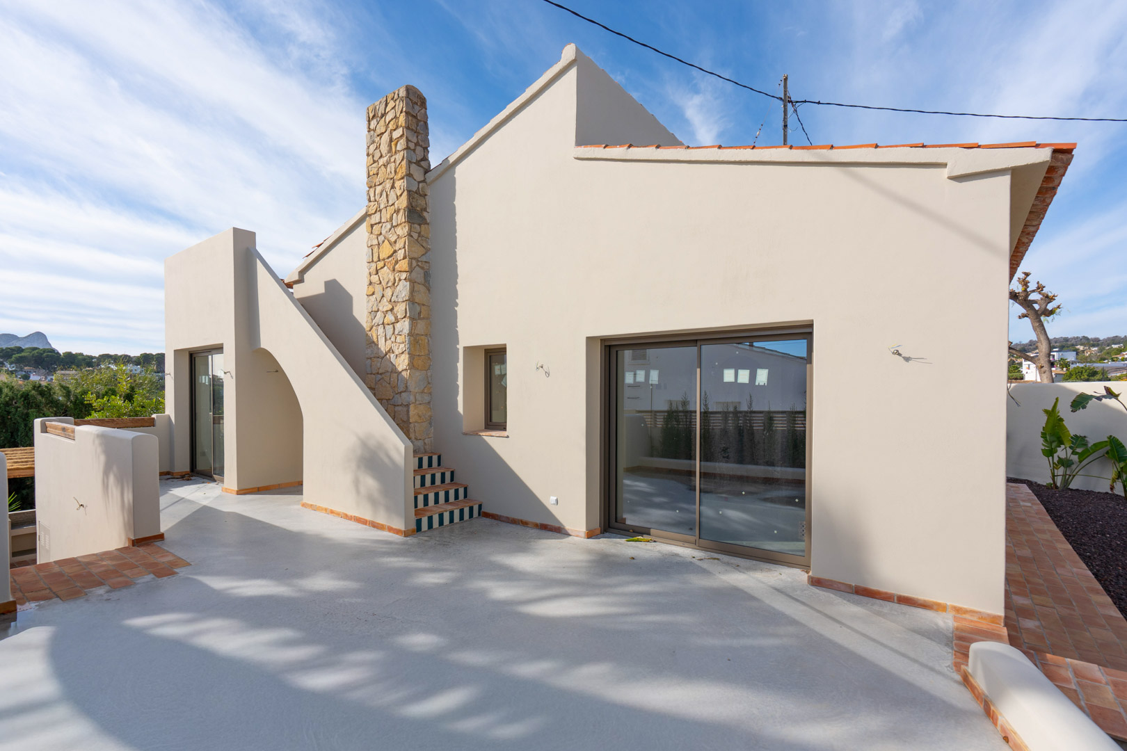 Exterior de la villa con amplia terraza de hormigón pulido, chimenea de piedra natural y escalera decorada con azulejos. Se aprecia el Peñón de Ifach al fondo.
