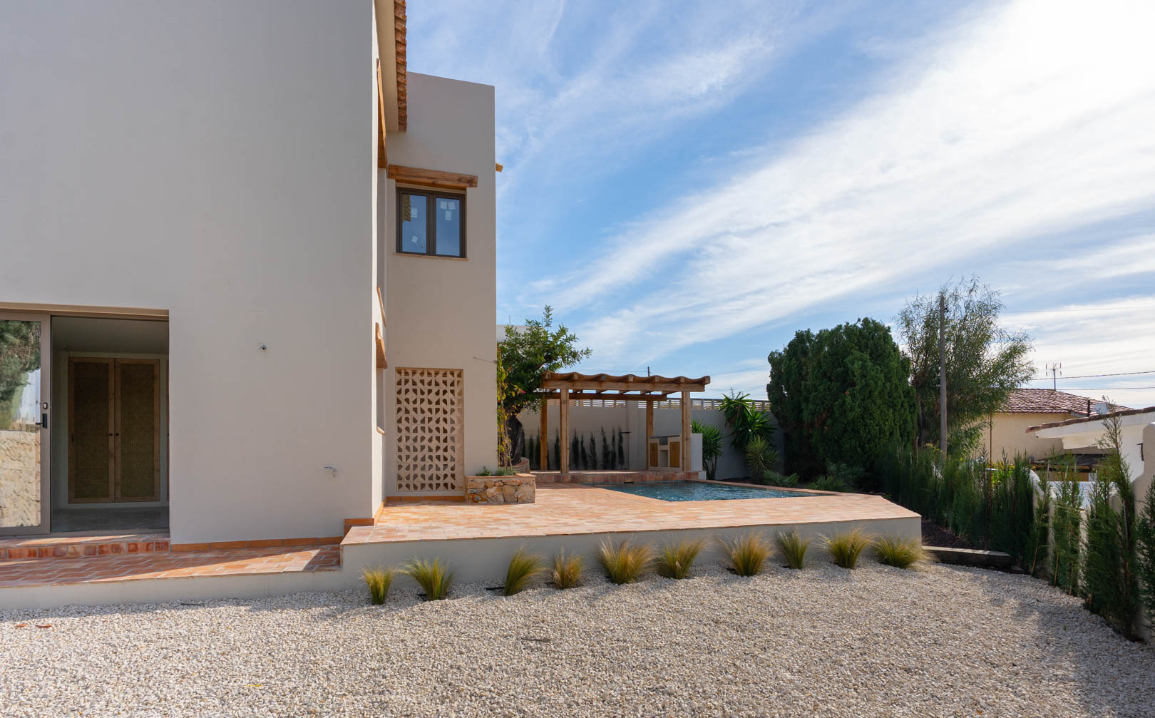 Exterior de la villa con terraza de terracota, piscina de nueva construcción y pérgola de madera sobre la zona de barbacoa, rodeado de un jardín de grava.
