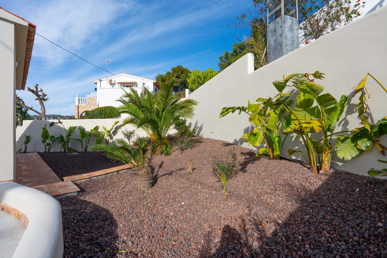 Jardín mediterráneo con grava decorativa, palmeras y bananos junto a un muro perimetral blanco y un sendero de baldosas de terracota.