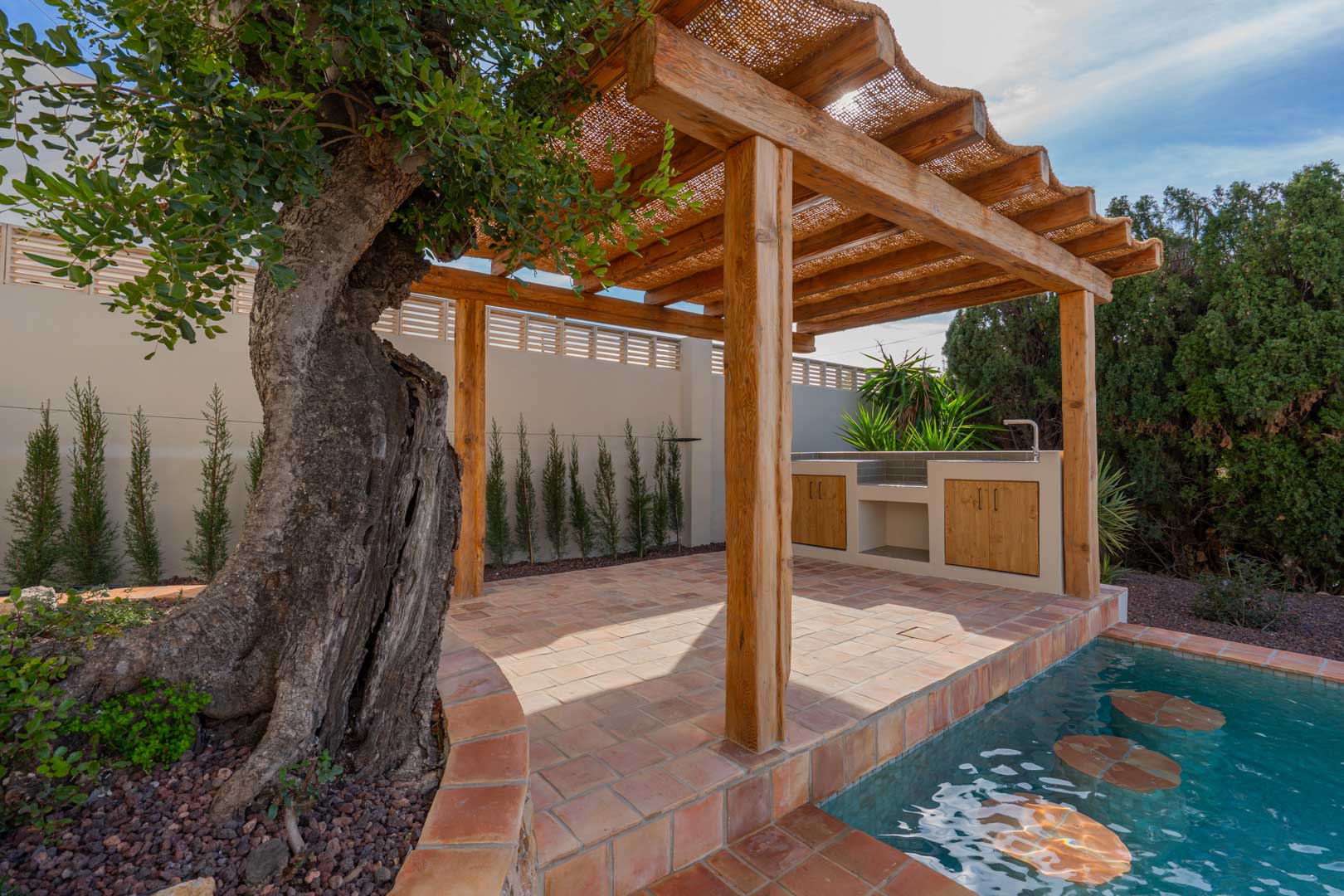 Pérgola de madera con cubierta de fibras naturales sobre suelo de terracota, junto a una piscina con taburetes integrados y cocina exterior equipada junto a un olivo.