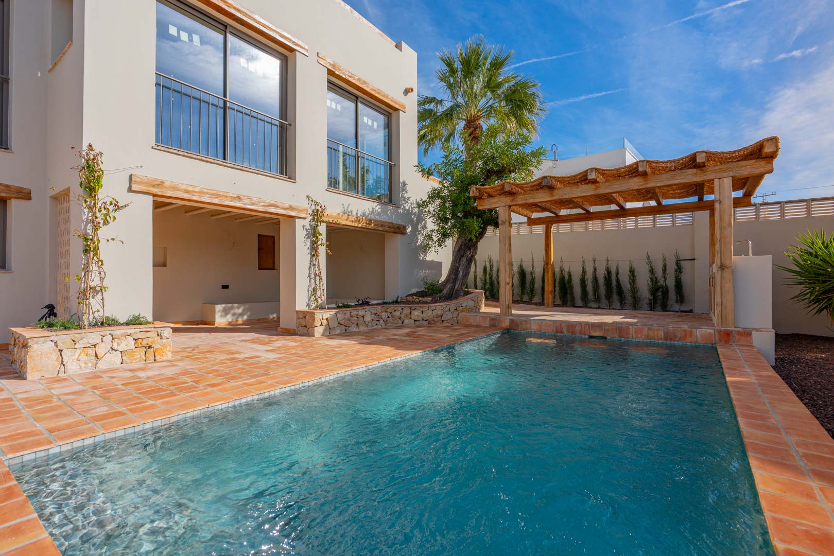 Piscina de nueva construcción rodeada de suelos de terracota, con una pérgola de madera y acabados en piedra natural junto a la fachada de la villa.