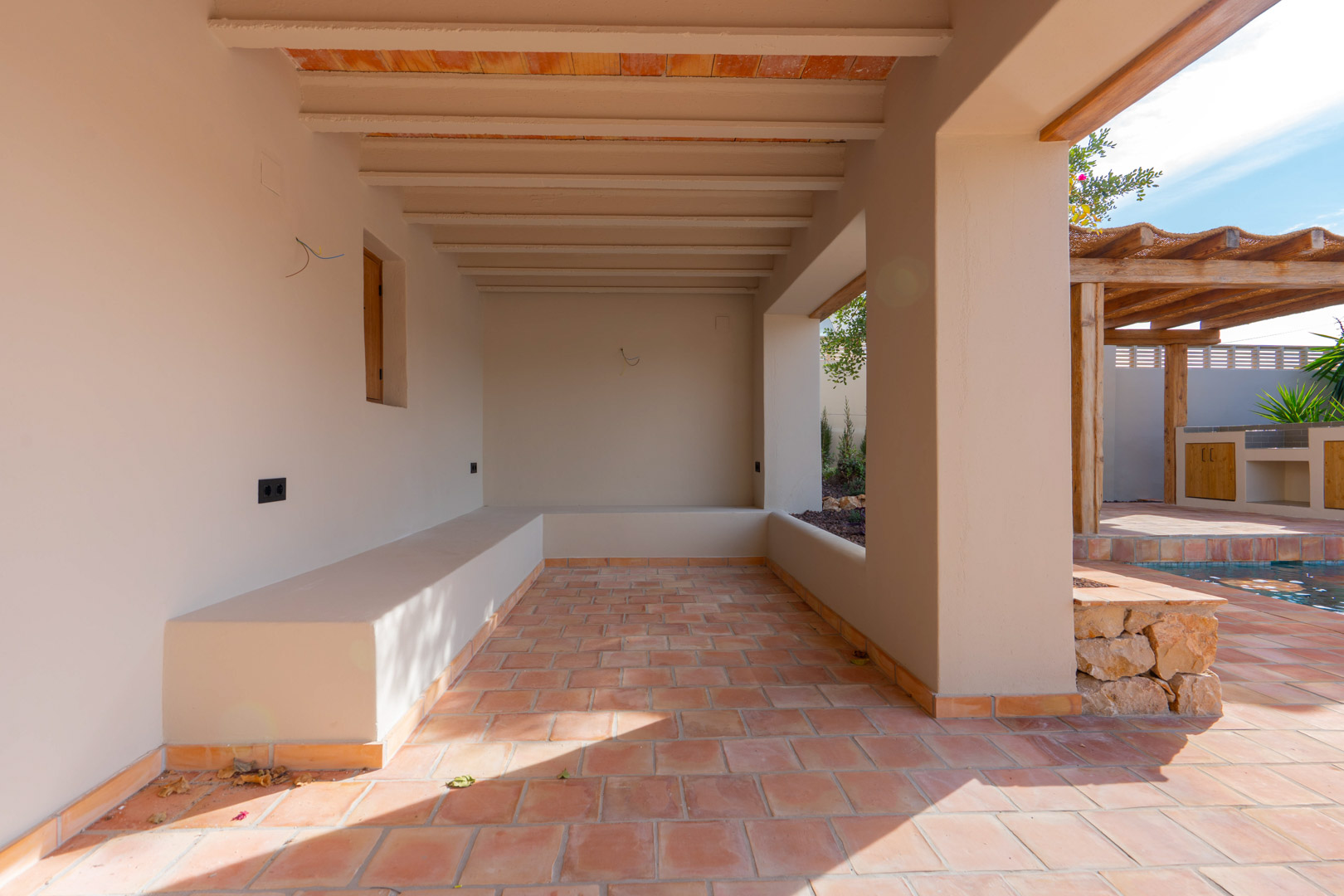 Porche cubierto con suelos de terracota, vigas vistas y bancos de obra integrados, con vistas a la zona de piscina y pérgola exterior.