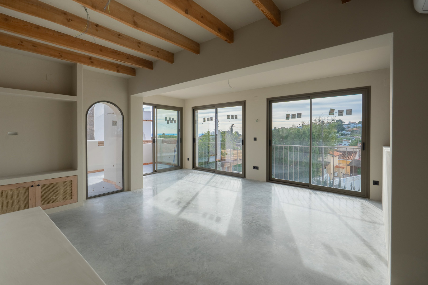 Salón luminoso con vigas de madera vista, suelo de hormigón pulido y amplios ventanales con vistas al mar. Incluye estanterías de obra con acabados en arcilla natural.