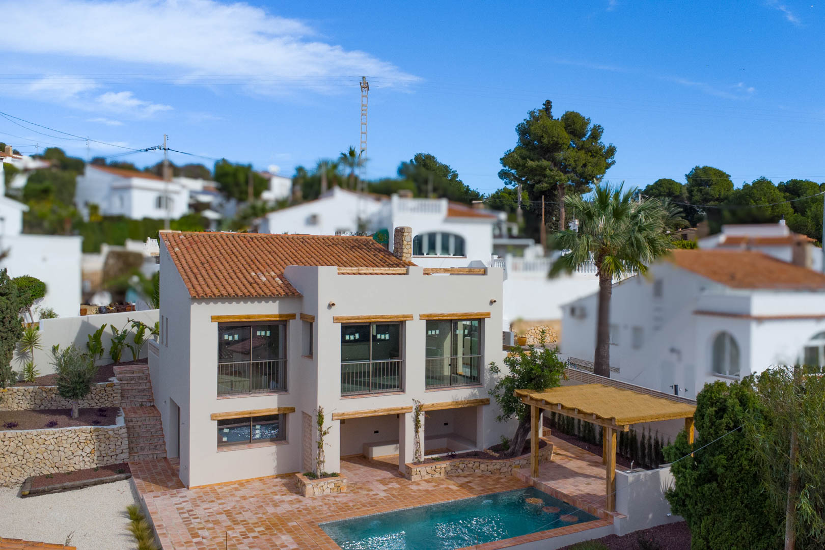 Vista exterior de la villa con fachada mediterránea, amplios ventanales, tejado de terracota y zona de piscina con pérgola de madera y muros de piedra natural.