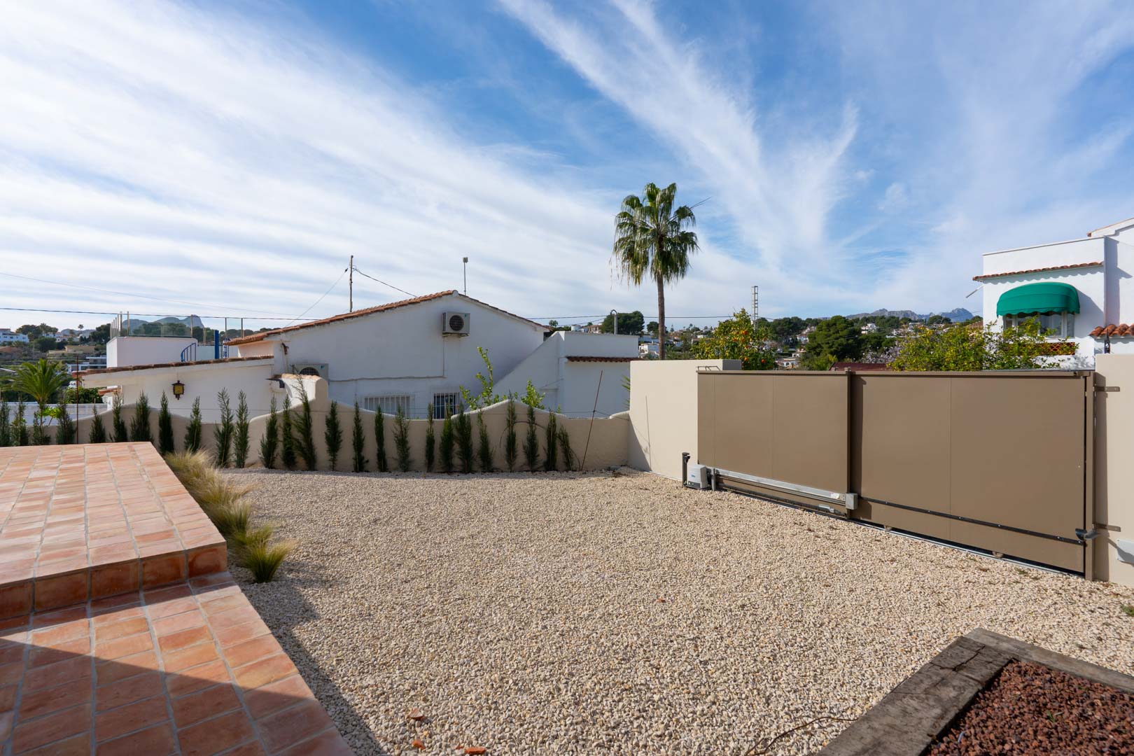 Zona de aparcamiento exterior con grava y puerta corredera motorizada, flanqueada por una terraza de terracota y vegetación mediterránea.