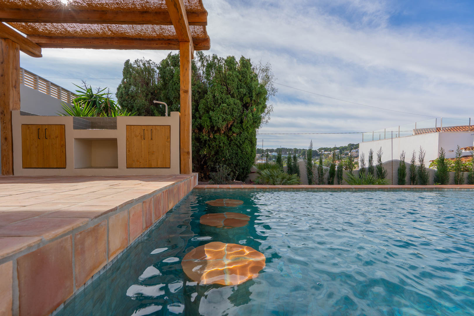Zona de piscina con taburetes sumergidos, barbacoa exterior bajo pérgola de madera y suelos de terracota tradicional con vistas despejadas.