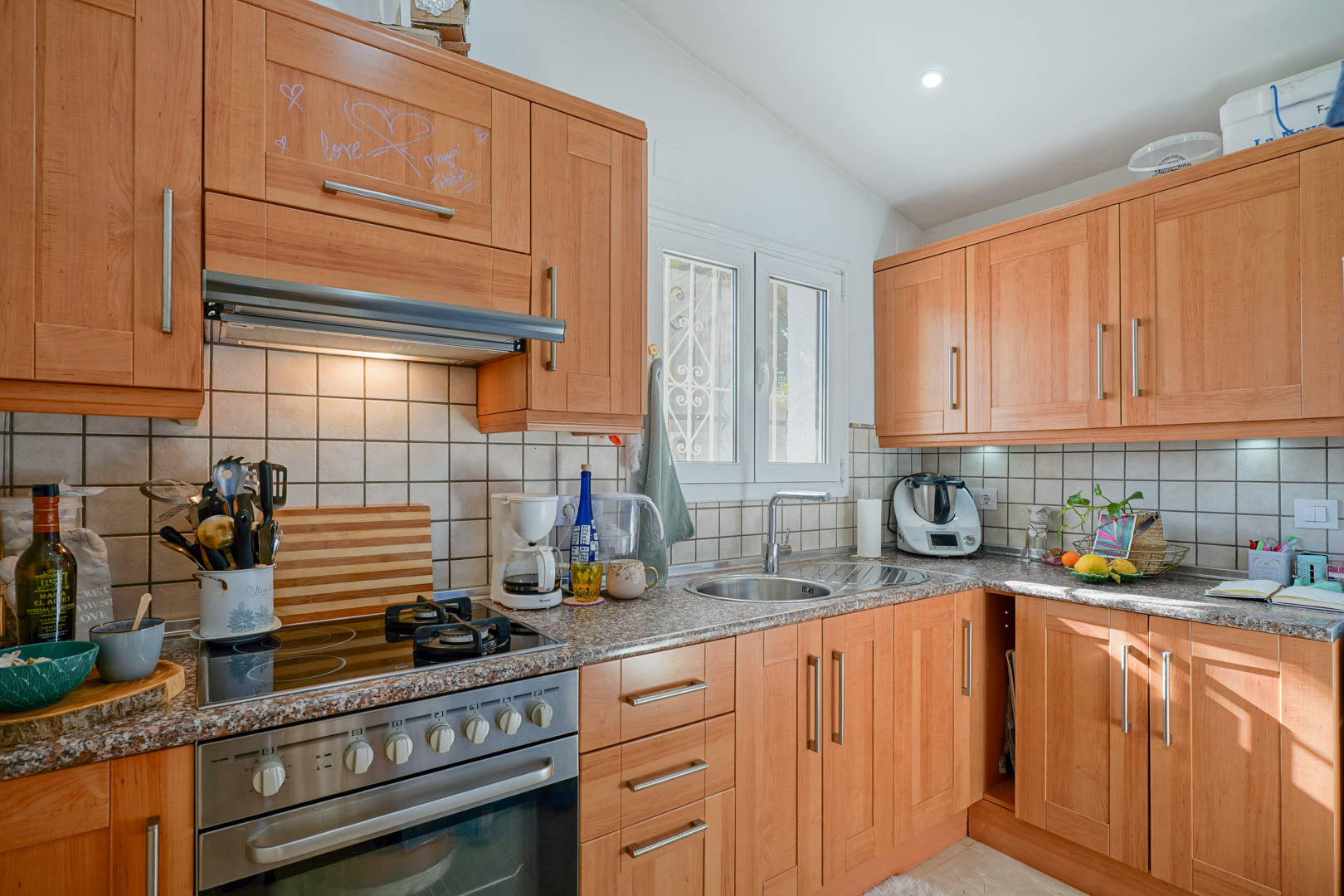 Cocina independiente con mobiliario de madera, encimera de granito y placa mixta. Incluye fregadero circular de acero inoxidable y ventana al exterior con luz natural.
