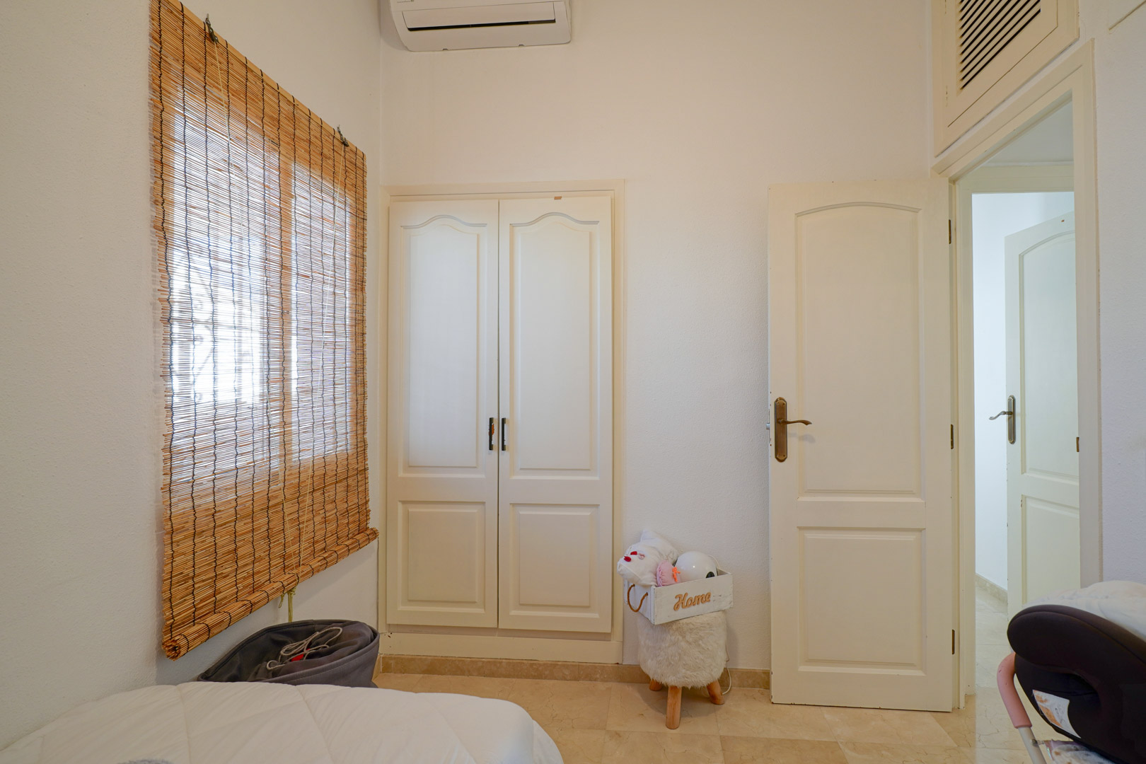 Dormitorio luminoso con armario empotrado blanco, aire acondicionado individual y ventana con estor de bambú sobre suelos de gres.
