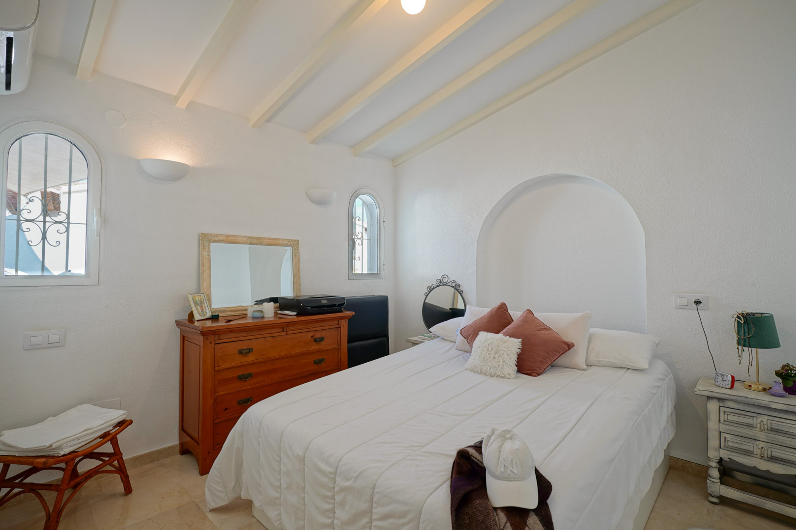 Dormitorio luminoso con techos de vigas blancas, ventanas de arco y una hornacina decorativa sobre la cama. Incluye aire acondicionado y suelos de mármol.