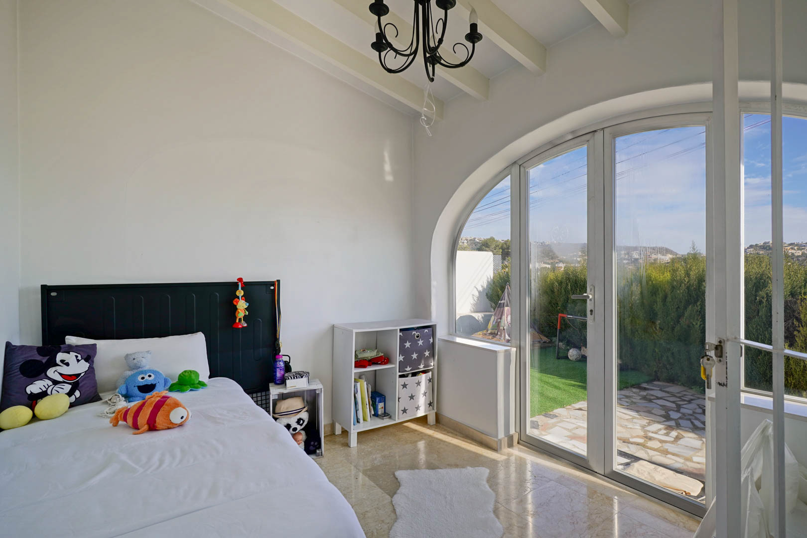 Dormitorio luminoso con techos de vigas blancas y un gran ventanal en arco que ofrece acceso directo al jardín y abundante luz natural sobre el suelo de mármol.