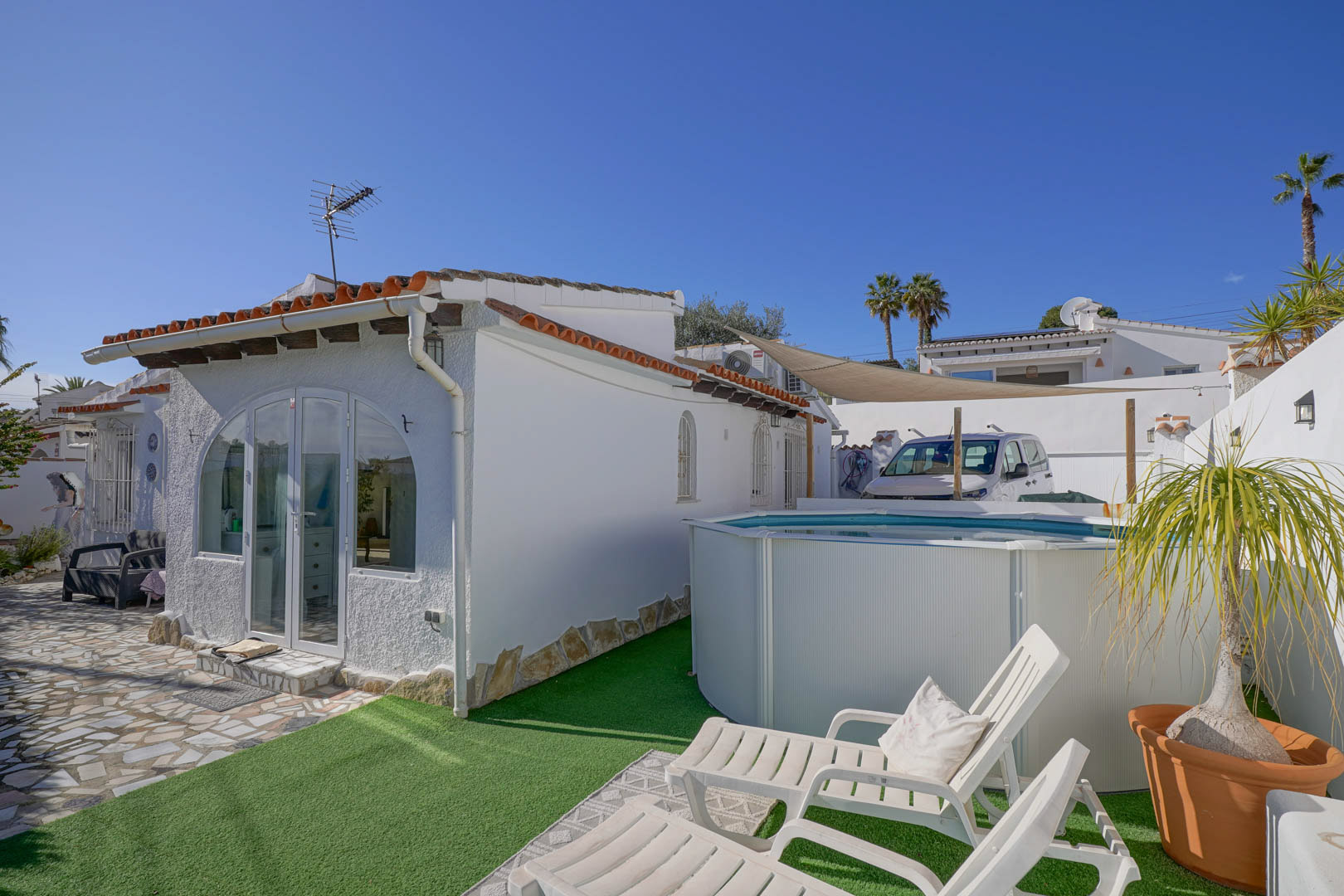 Exterior de la villa con fachada blanca, porche acristalado y zona de ocio que incluye una piscina portátil sobre césped artificial y tumbonas blancas.