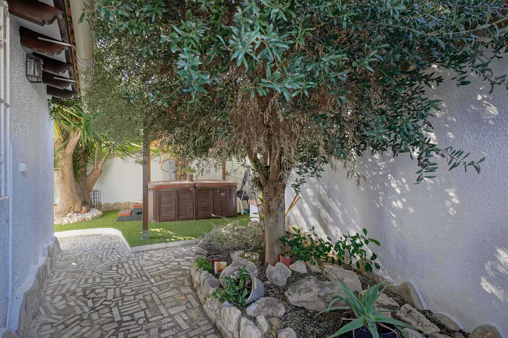 Terraza exterior con jacuzzi privado sobre césped artificial, rodeada de vegetación mediterránea y un camino de piedra natural junto a muros blancos.