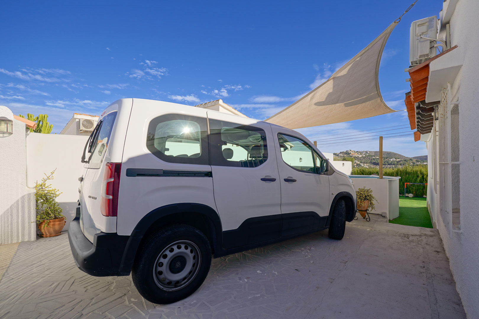 Zona de aparcamiento privado con pavimento impreso y toldo vela para sombra, situada junto a la fachada blanca de la villa con vistas despejadas al entorno.