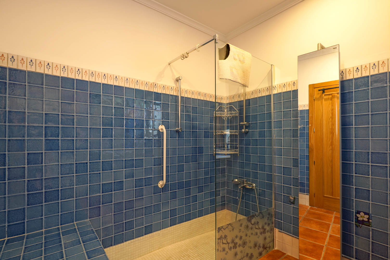 Baño con ducha de obra revestida en azulejos azules, mampara de cristal con detalle floral, suelo de mosaico blanco y asidero de seguridad para mayor accesibilidad.