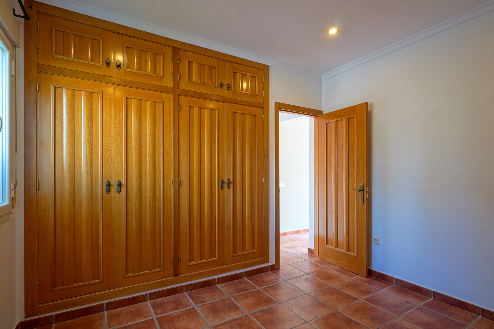 Dormitorio con amplio armario empotrado de madera de suelo a techo, suelos de gres rústico y paredes blancas con iluminación empotrada.
