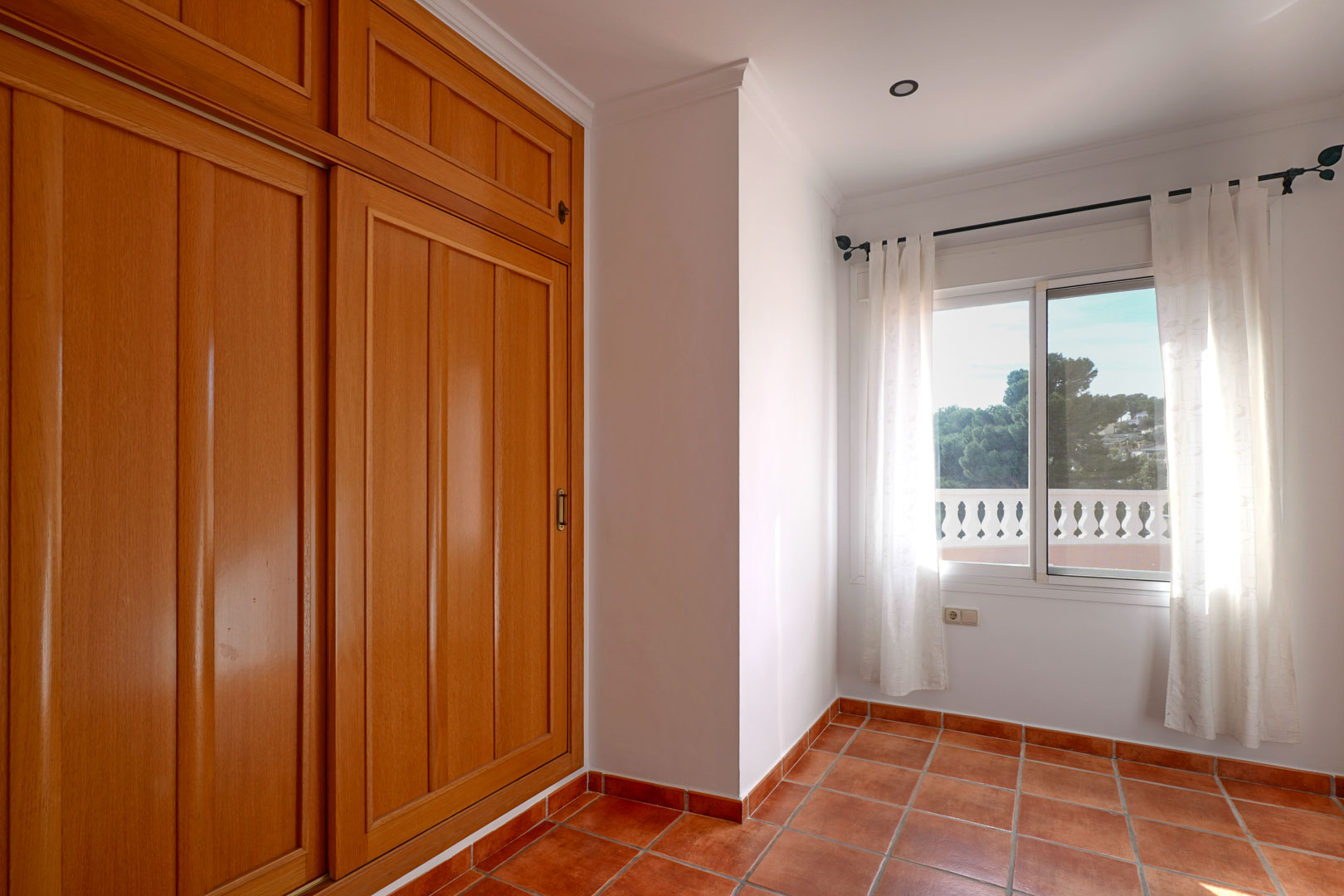 Dormitorio con amplio armario empotrado de madera, suelos de gres rústico y ventana con luz natural que muestra la balaustrada de la terraza.
