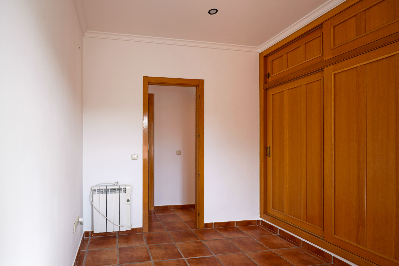 Dormitorio con armario empotrado de madera, suelos de gres rústico y radiador eléctrico. La estancia presenta paredes blancas, molduras en el techo y acceso al pasillo.