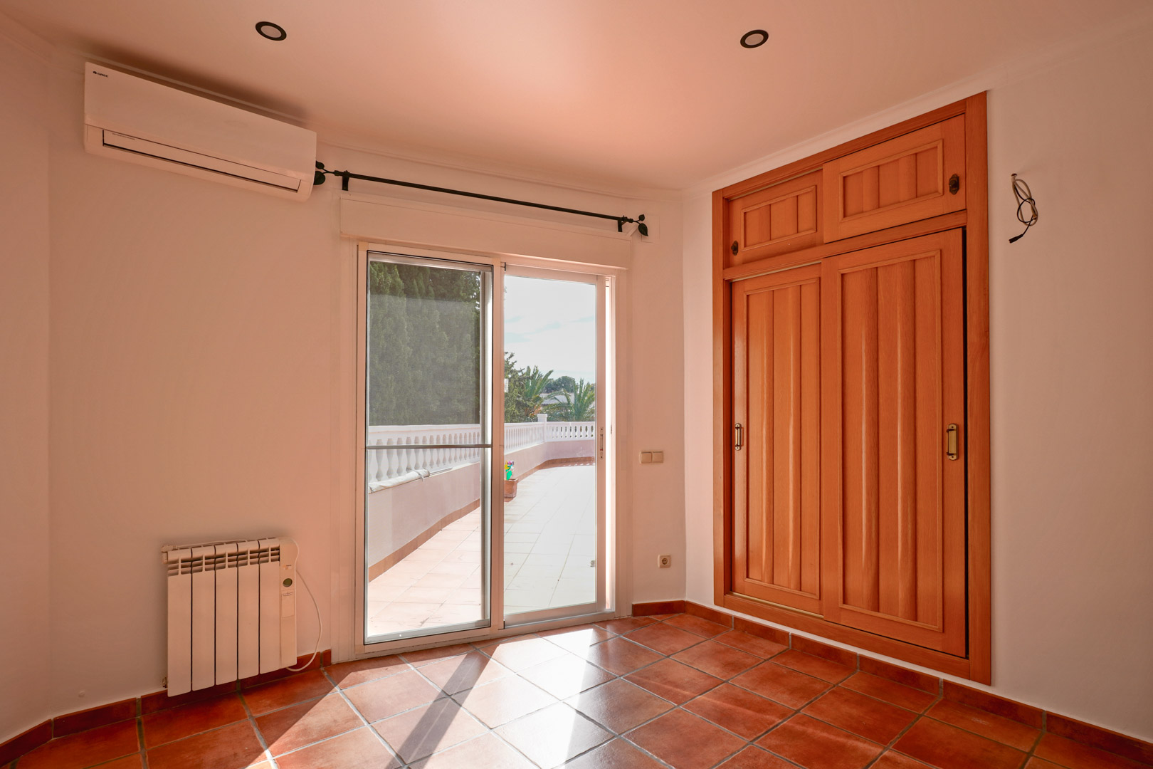 Dormitorio con armario empotrado de madera, suelos de terracota y acceso directo a la terraza. Incluye aire acondicionado, radiador eléctrico e iluminación empotrada.