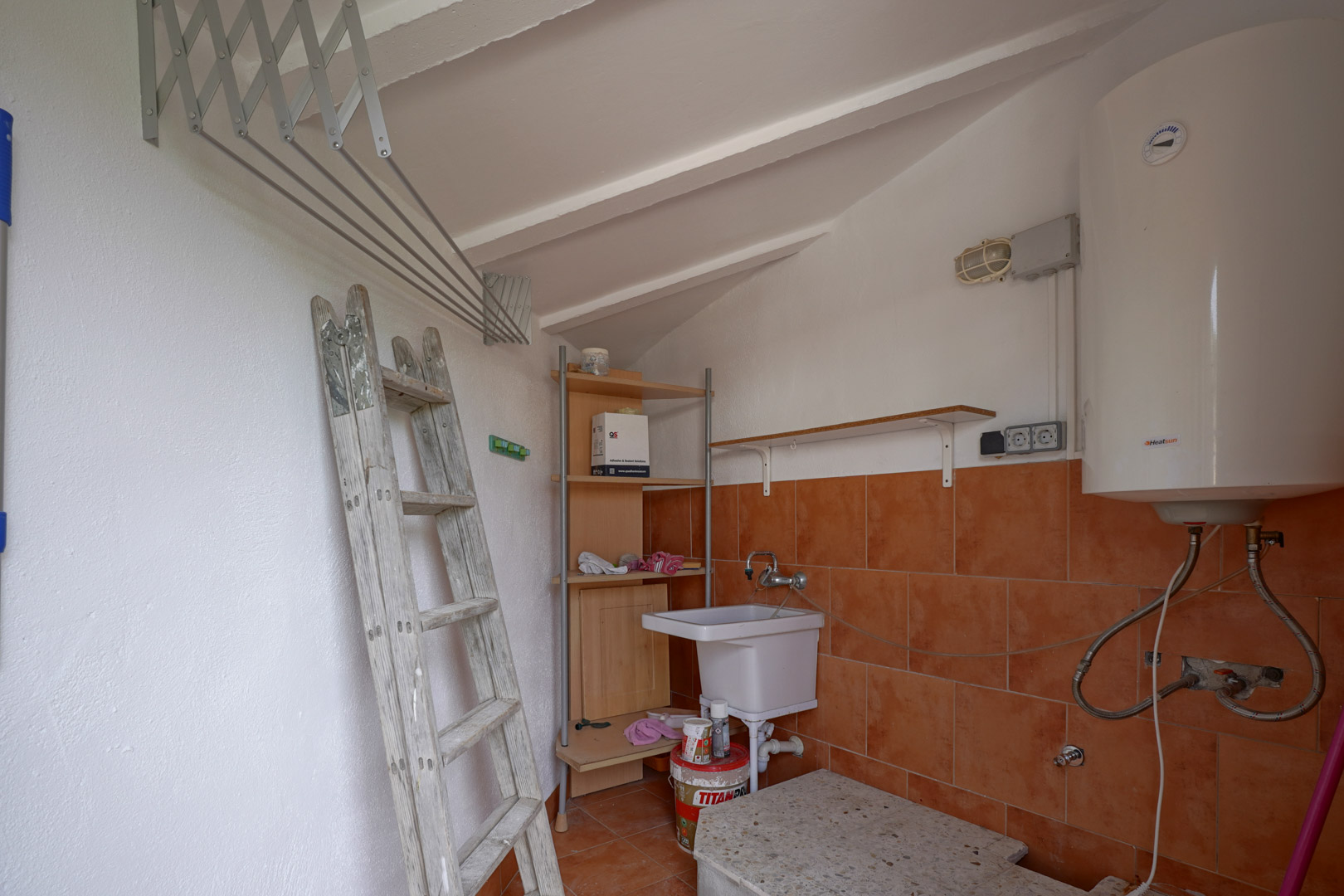 Lavadero y trastero funcional con techo inclinado, equipado con termo eléctrico, pila de lavado, estanterías y tendedero extensible de pared.
