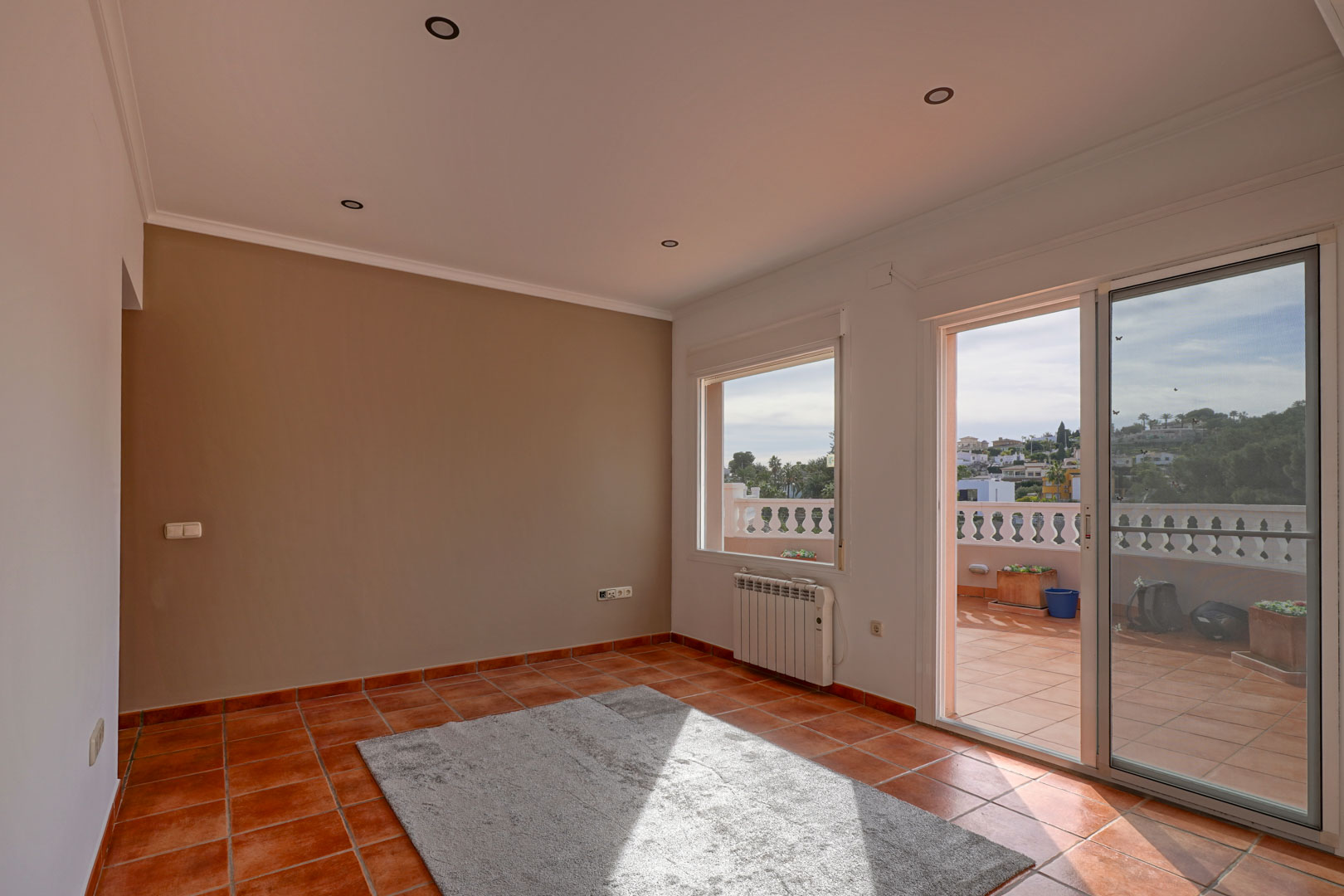 Salón con suelos de terracota, iluminación empotrada y acceso directo a una amplia terraza con balustrada. Incluye radiador eléctrico y pared de acento.