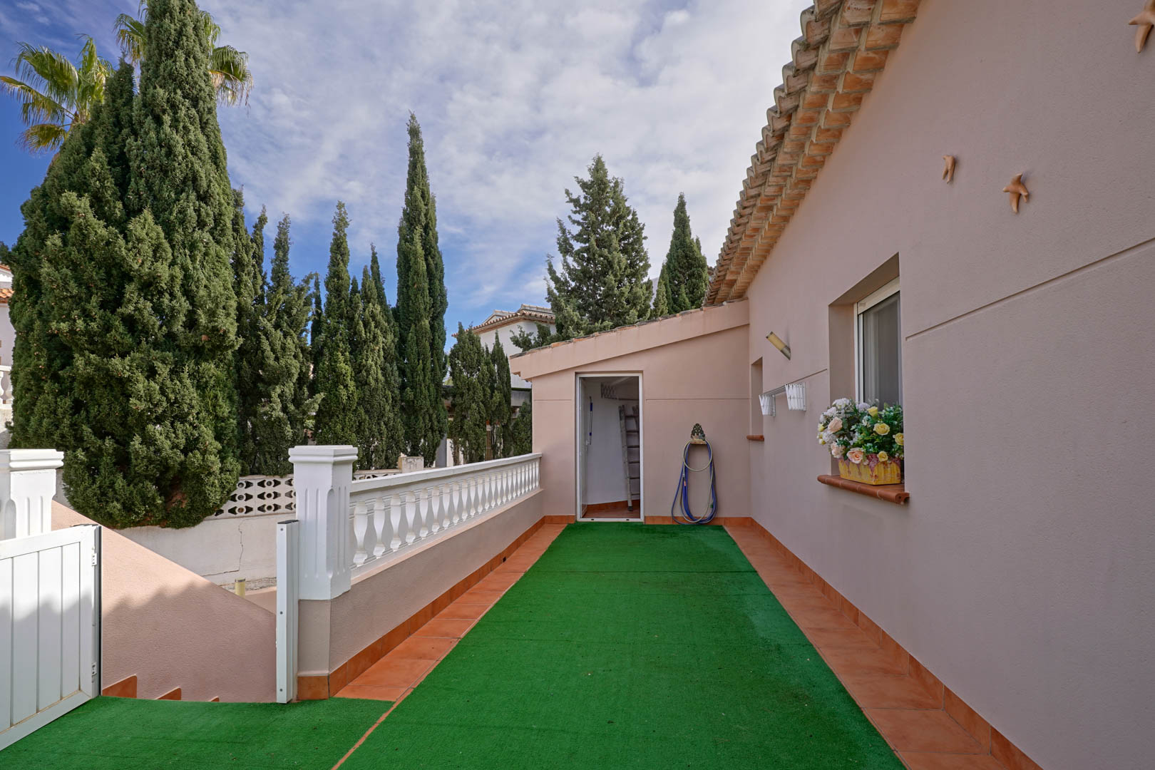 Terraza de acceso con césped artificial y balaustrada blanca, que conecta la entrada principal con el lavadero bajo un entorno de cipreses.