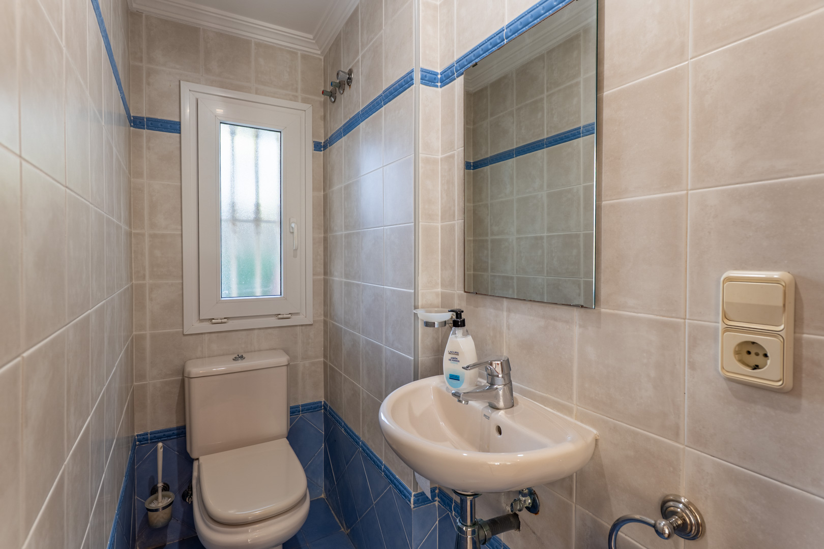 Aseo de cortesía con revestimiento cerámico en tonos beige y azul, equipado con sanitario blanco, lavabo compacto y ventana de ventilación natural.