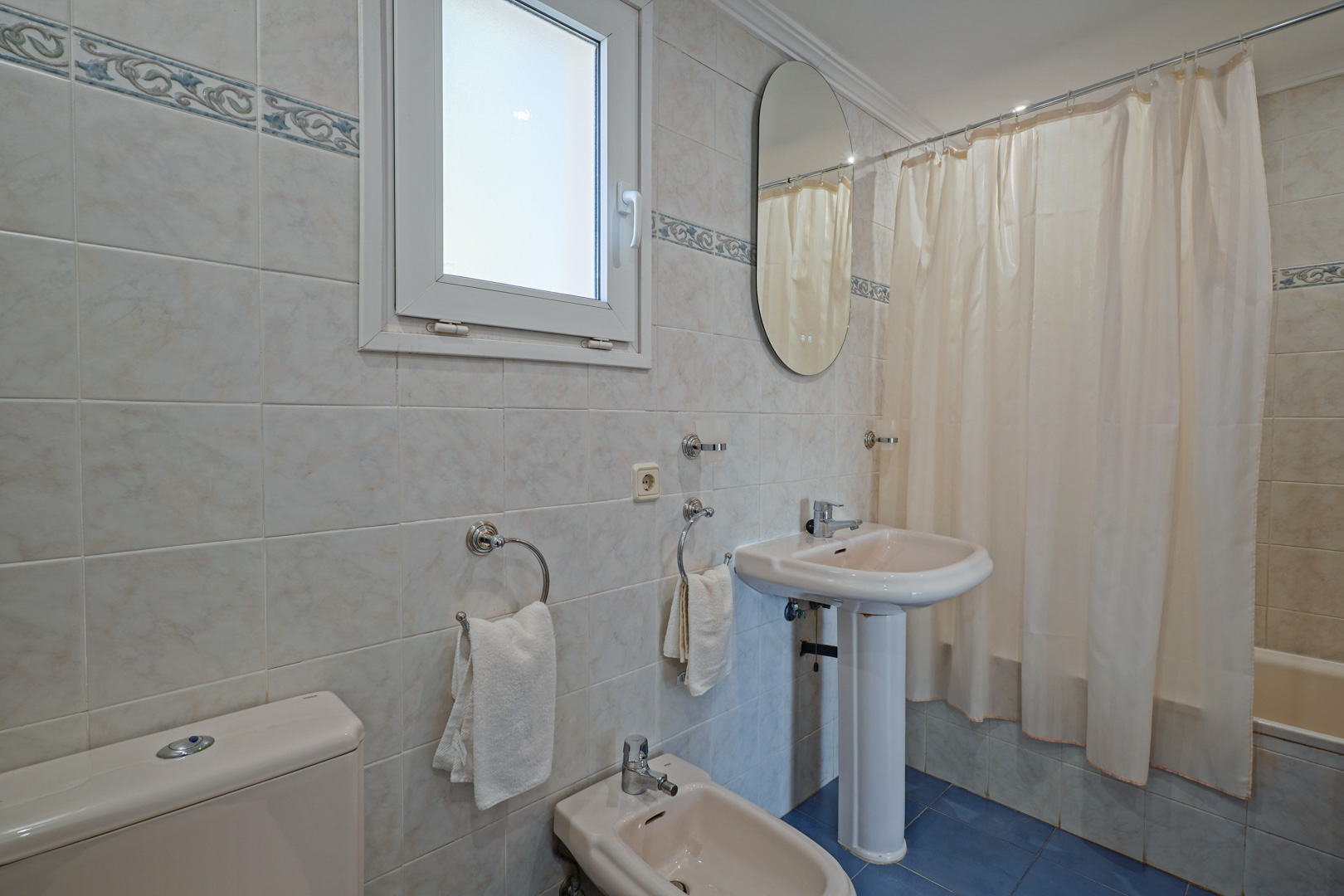 Baño familiar con sanitarios en tono crema, bidé, lavabo de pedestal y bañera. Presenta paredes alicatadas con cenefa decorativa, suelo de baldosa azul y ventana oscilobatiente.