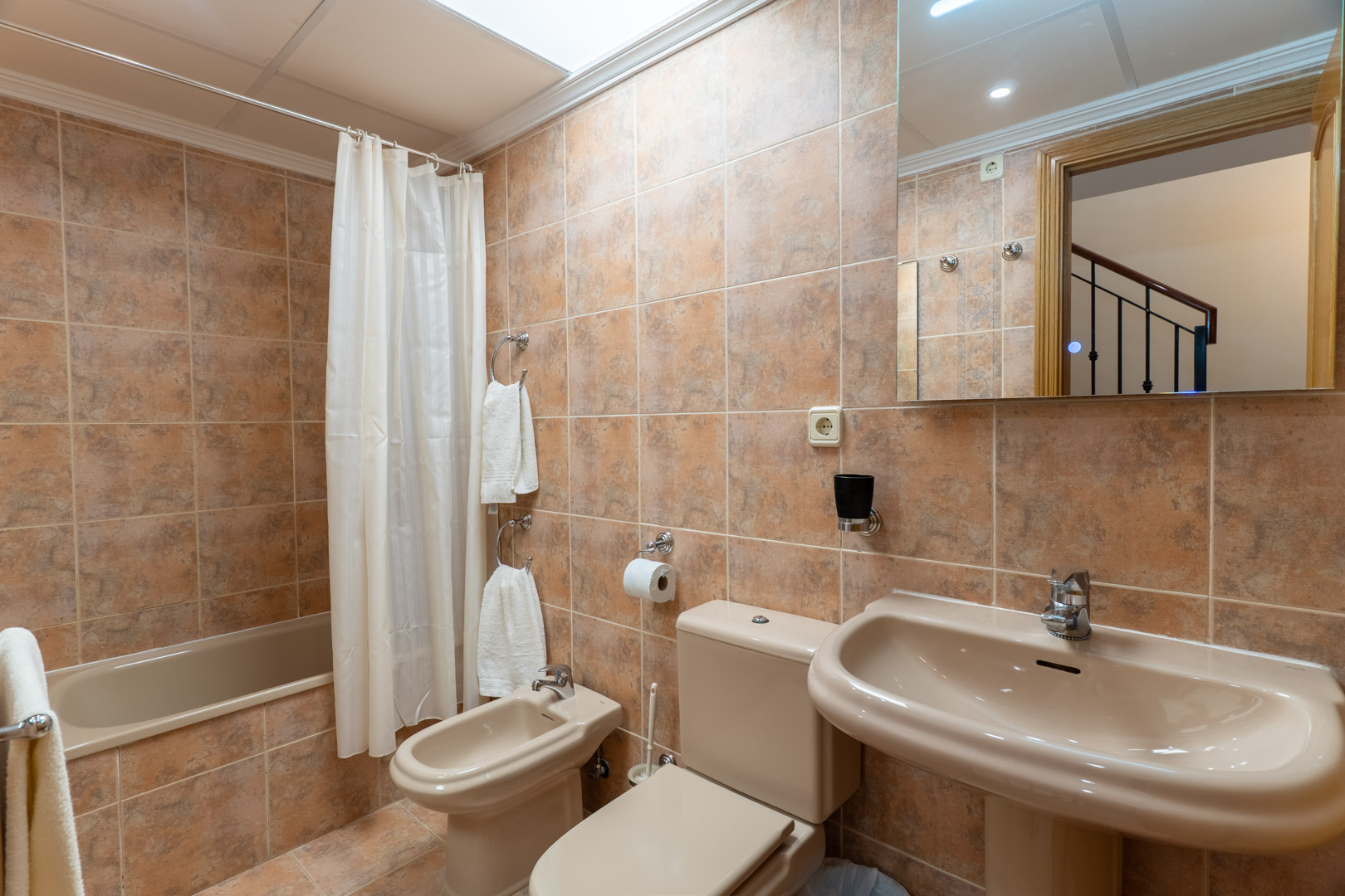 Cuarto de baño completo con bañera, bidé y lavabo de pedestal, revestido con azulejos cerámicos en tonos tierra y luz natural cenital.
