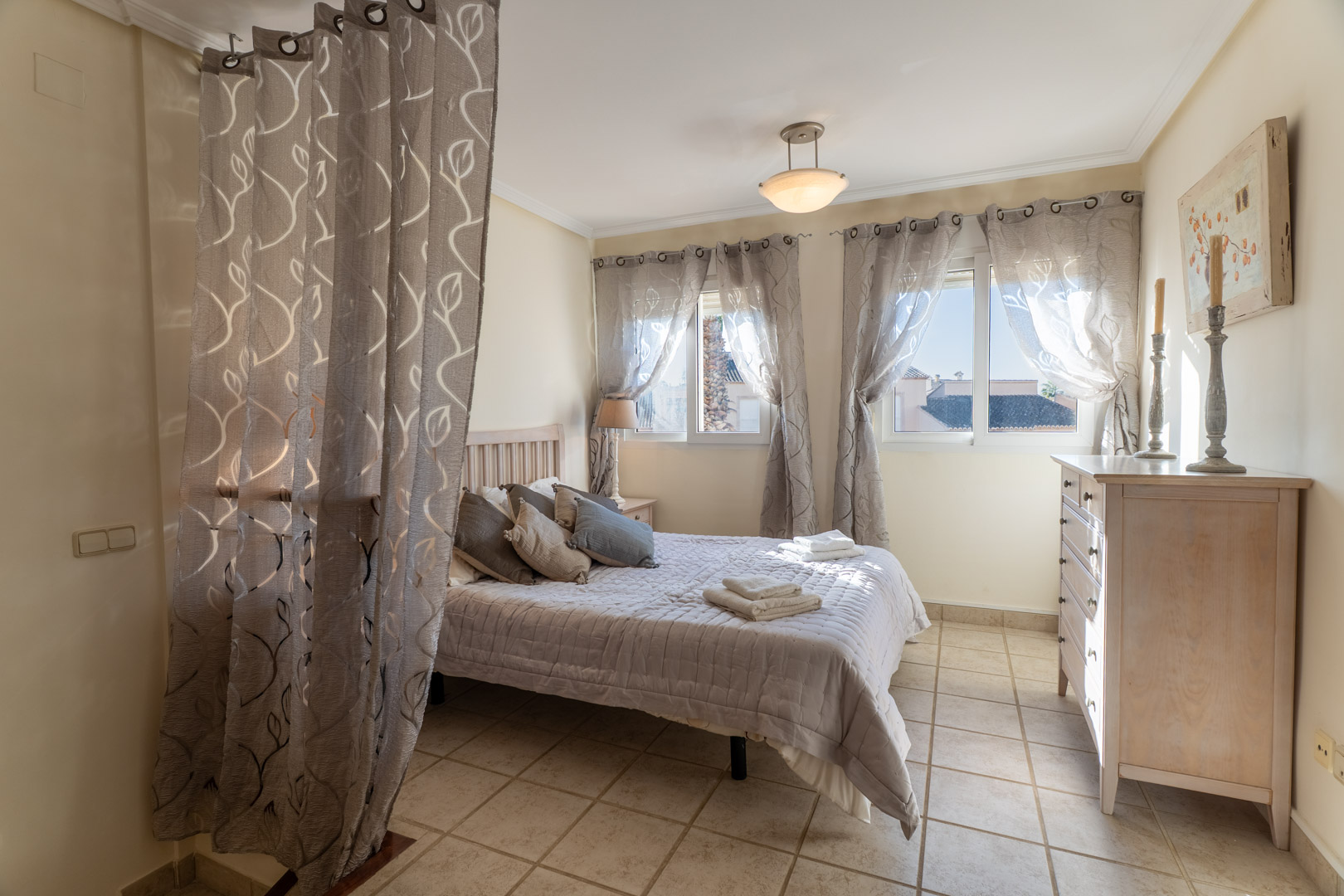Dormitorio con suelos de gres, cama doble, cómoda de madera y ventanas con cortinas estampadas que permiten la entrada de luz natural.