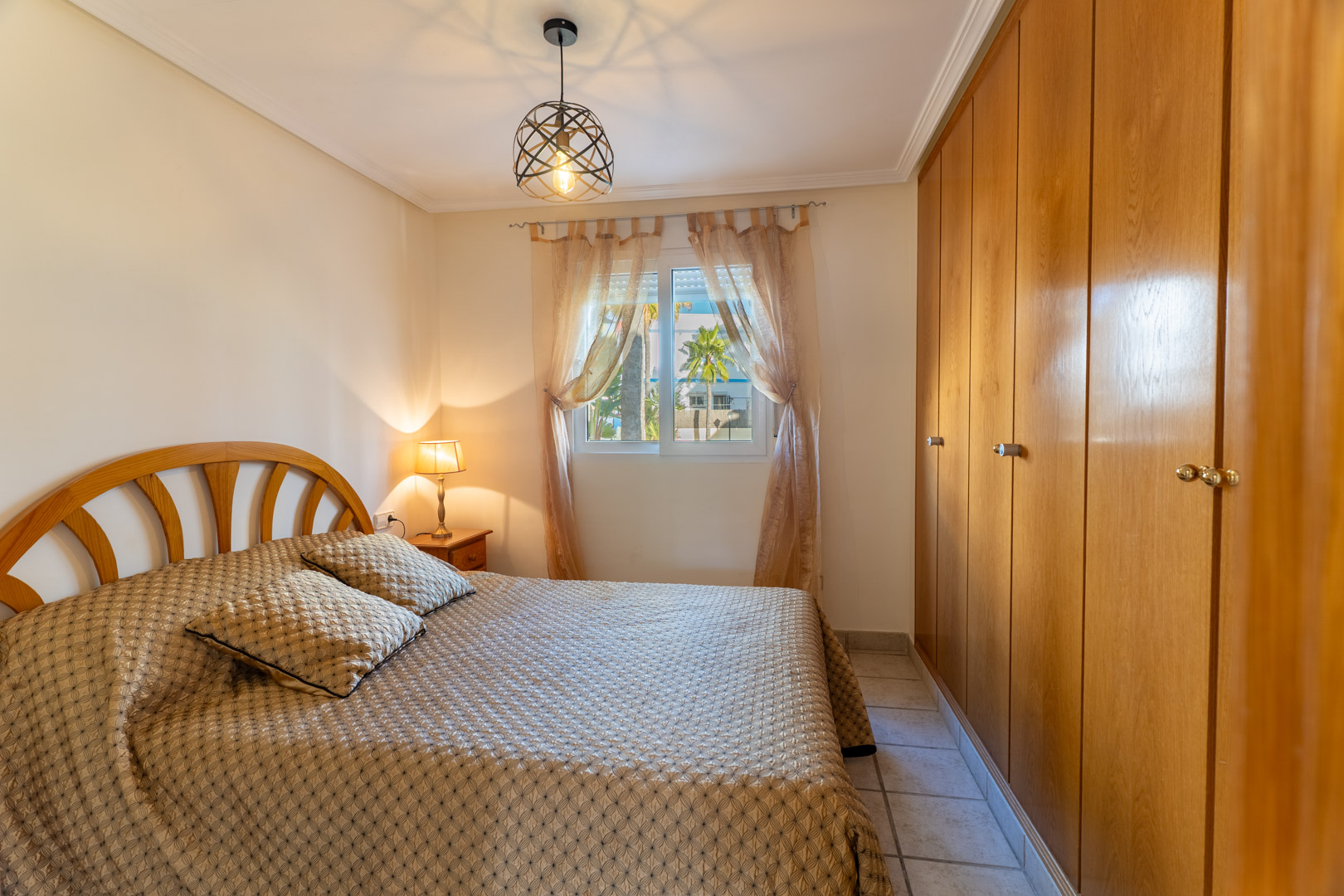 Dormitorio luminoso con amplios armarios empotrados de madera, cama doble y ventana con vistas a las zonas ajardinadas de la urbanización.