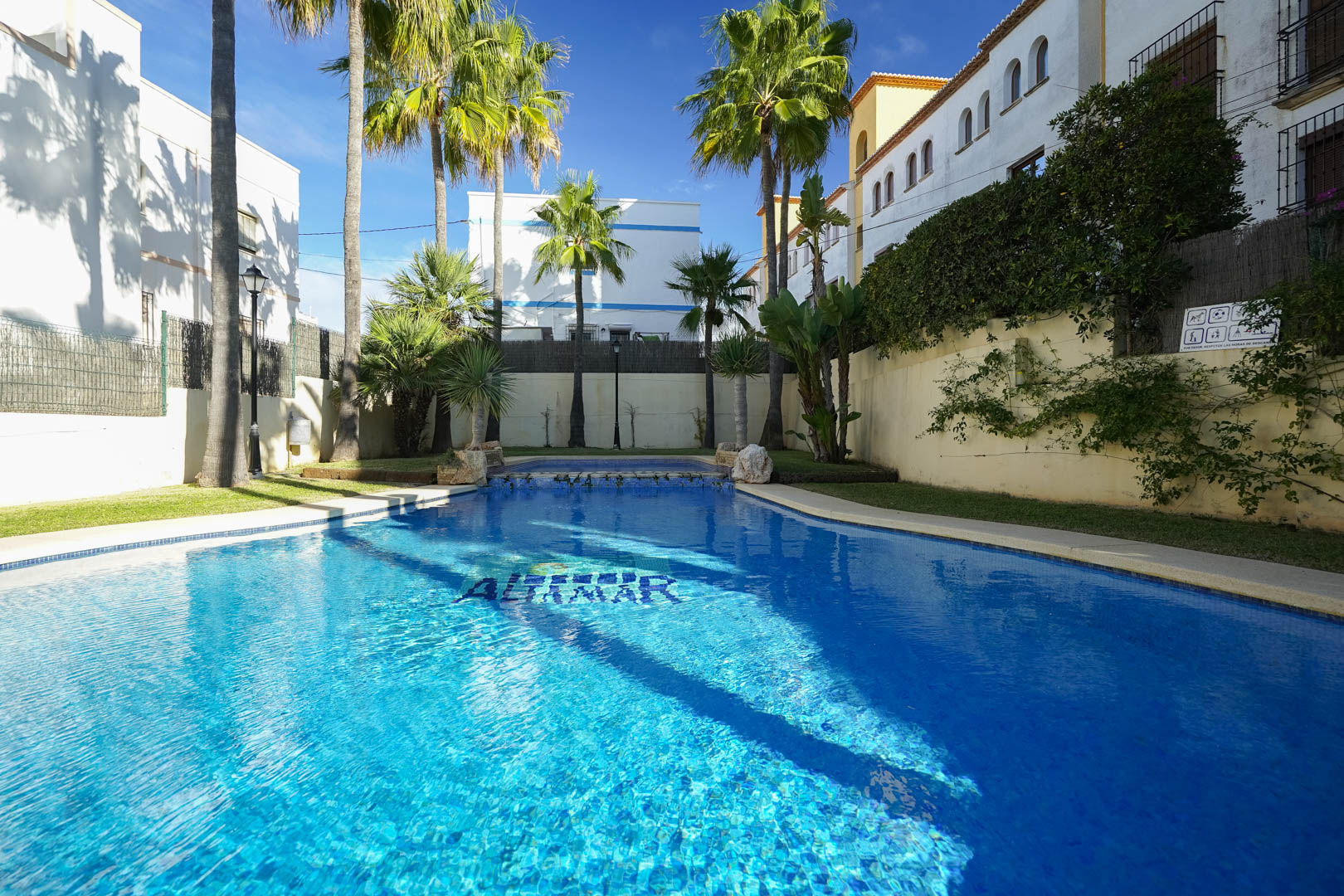 Piscina comunitaria con azulejos azules, rodeada de palmeras y zonas verdes cuidadas. El entorno ofrece privacidad y sol directo en una urbanización exclusiva de Jávea.