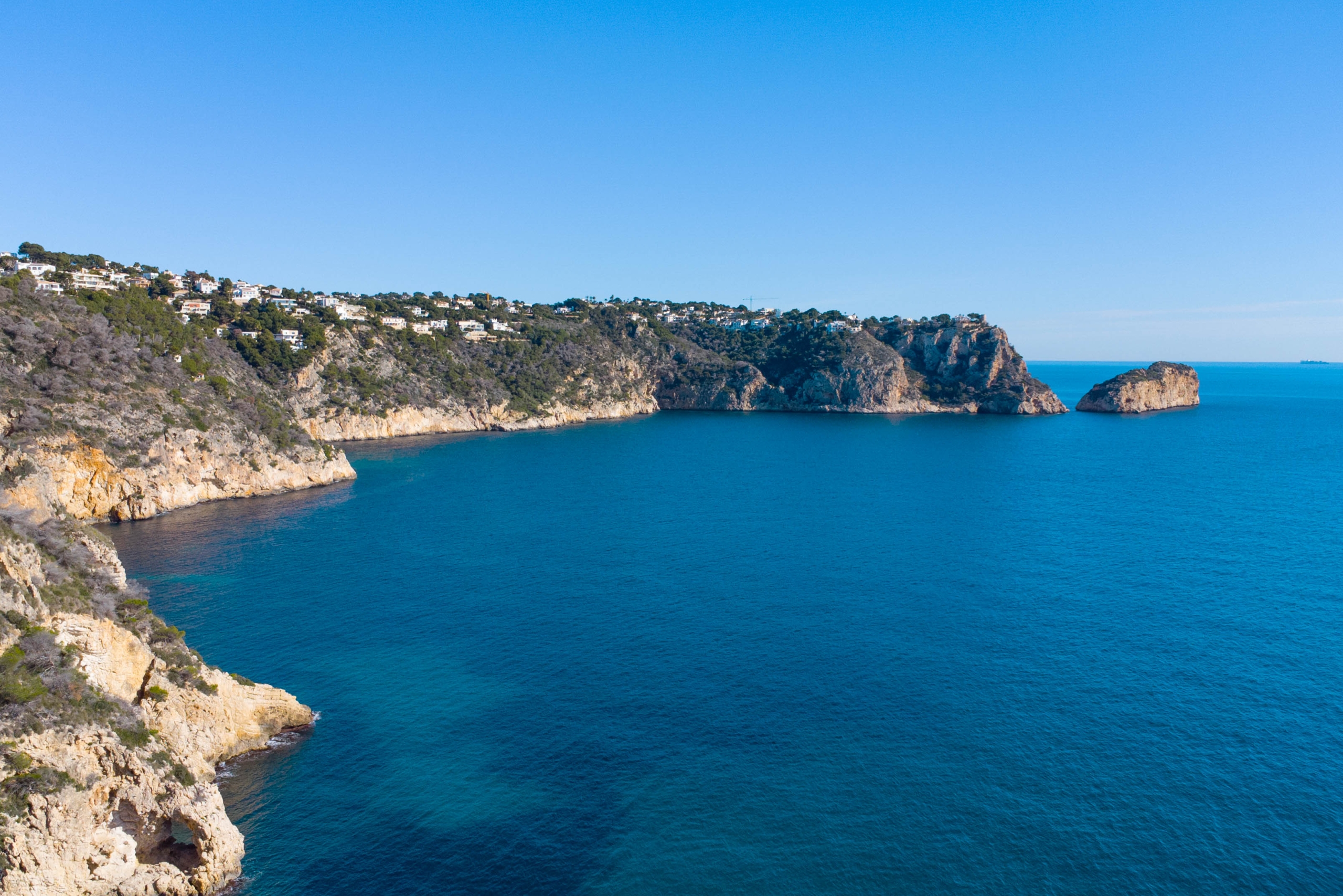 Vista aérea de los acantilados de Jávea con villas residenciales integradas en el paisaje costero y aguas cristalinas del Mediterráneo bajo un cielo despejado.