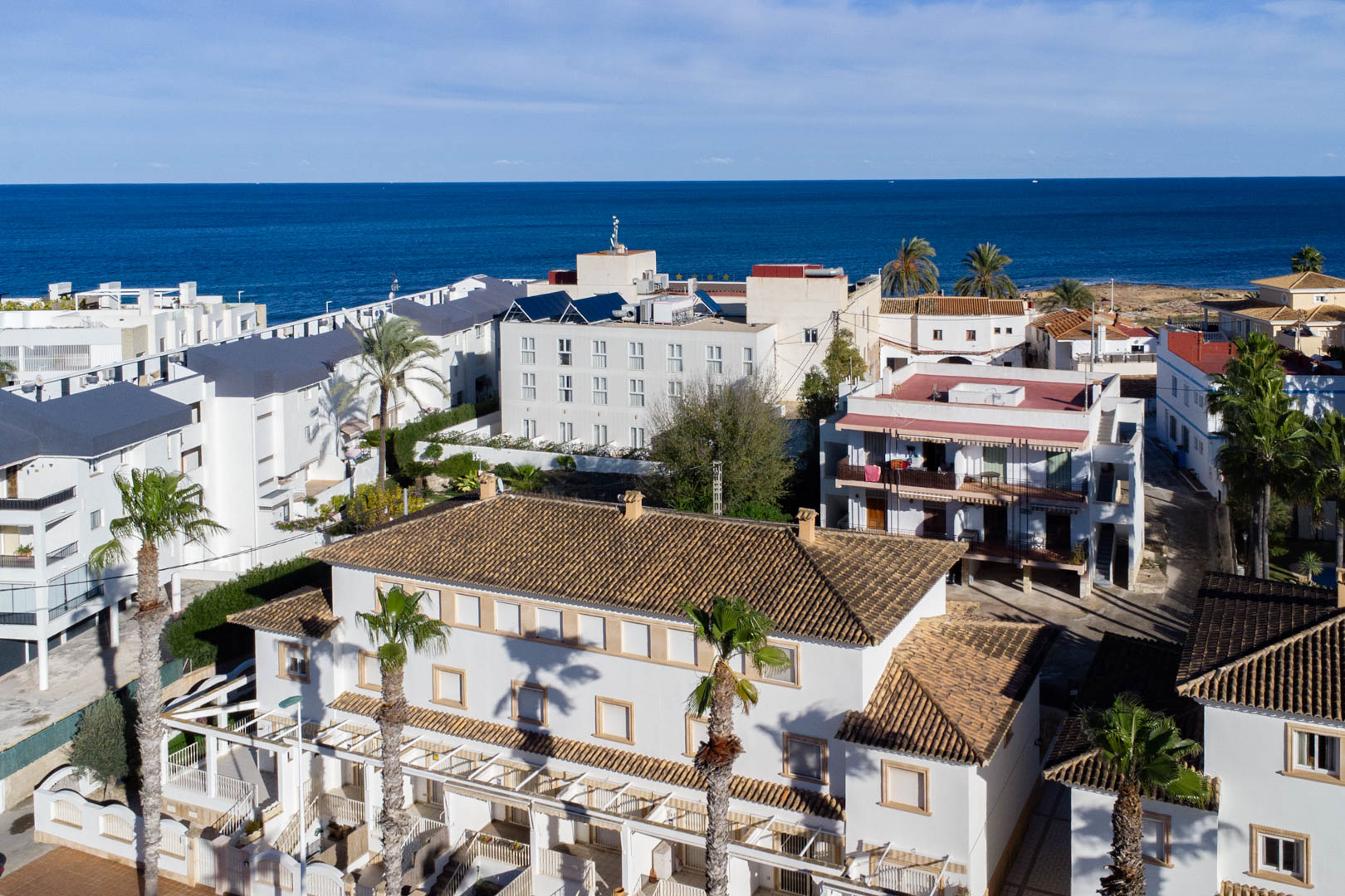 Vista aérea del complejo residencial con tejados de teja cerámica y fachadas blancas, destacando su proximidad al mar Mediterráneo y las palmeras circundantes.