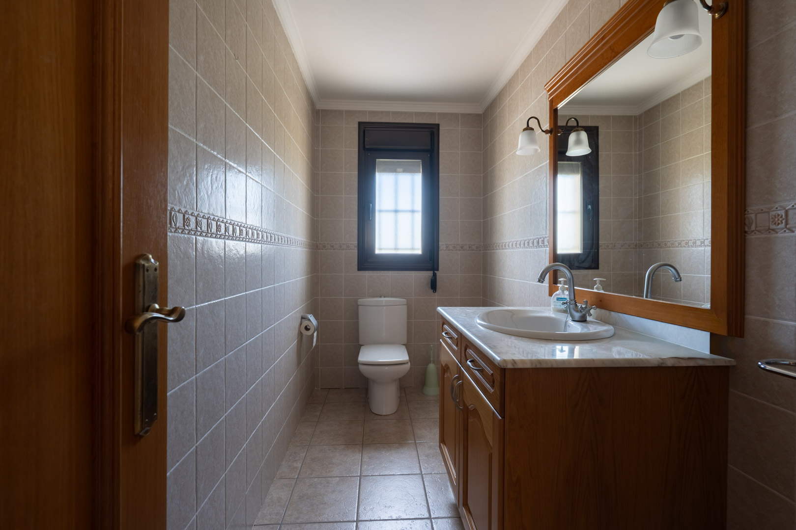 Baño con mueble de madera, encimera de mármol y espejo de marco ancho. Paredes revestidas con azulejos beige y cenefa decorativa bajo luz natural.