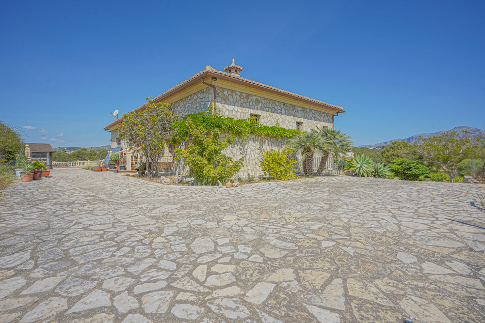 Fachada de piedra natural con amplio camino pavimentado, vegetación mediterránea, palmeras y tejado de teja cerámica bajo un cielo despejado.
